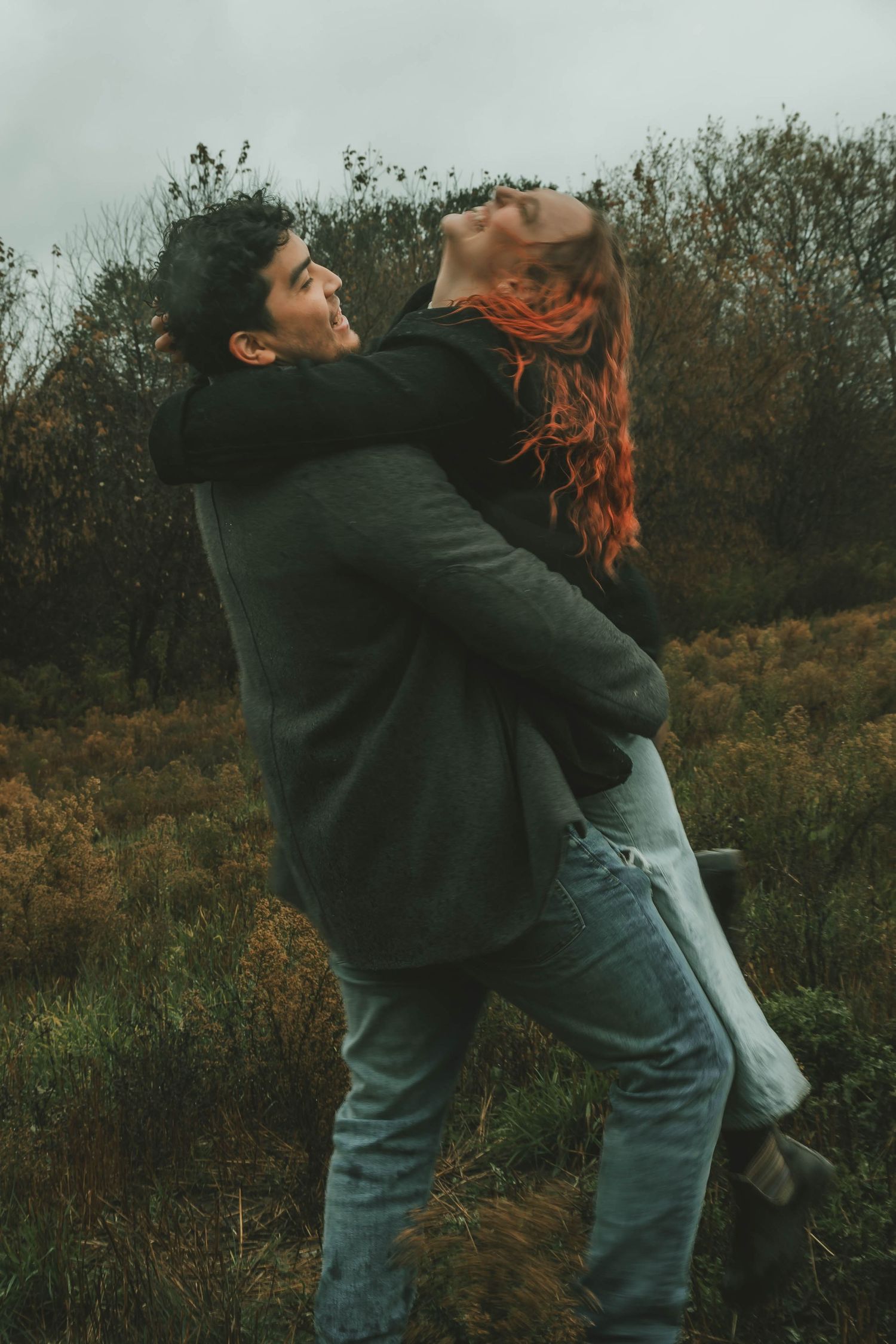 Playful couple dancing and twirling together in autumn field during overcast weather.