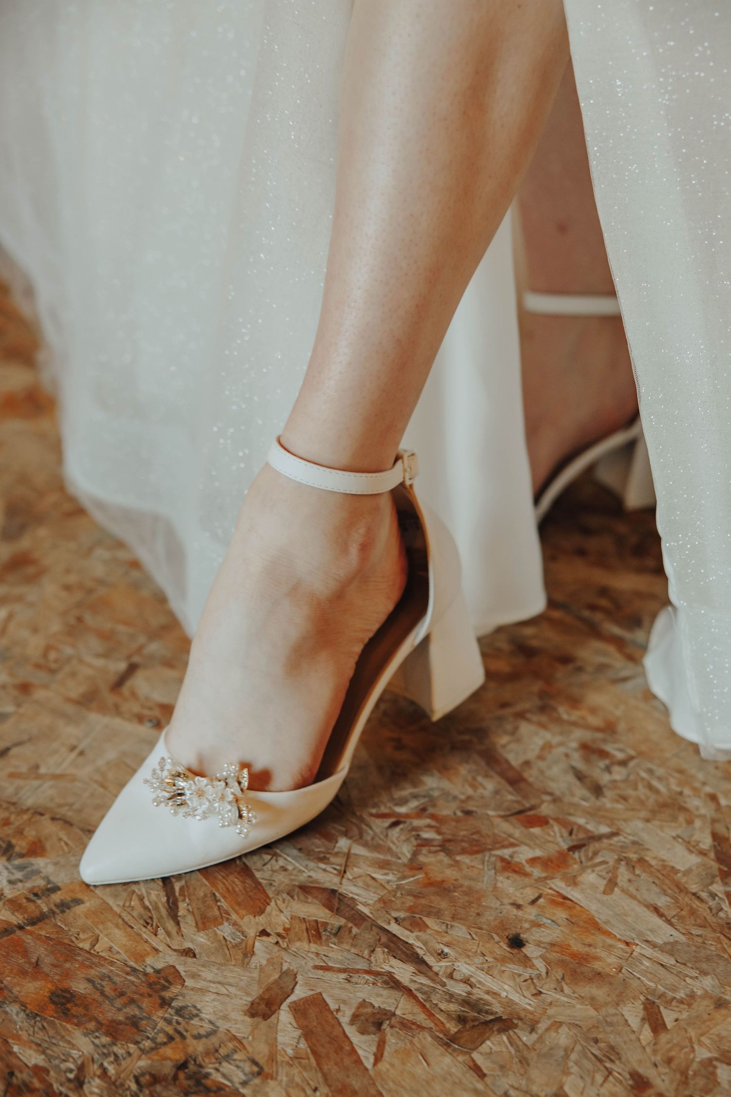 Close-up of elegant white bridal shoes with crystal embellishments on wooden flooring.