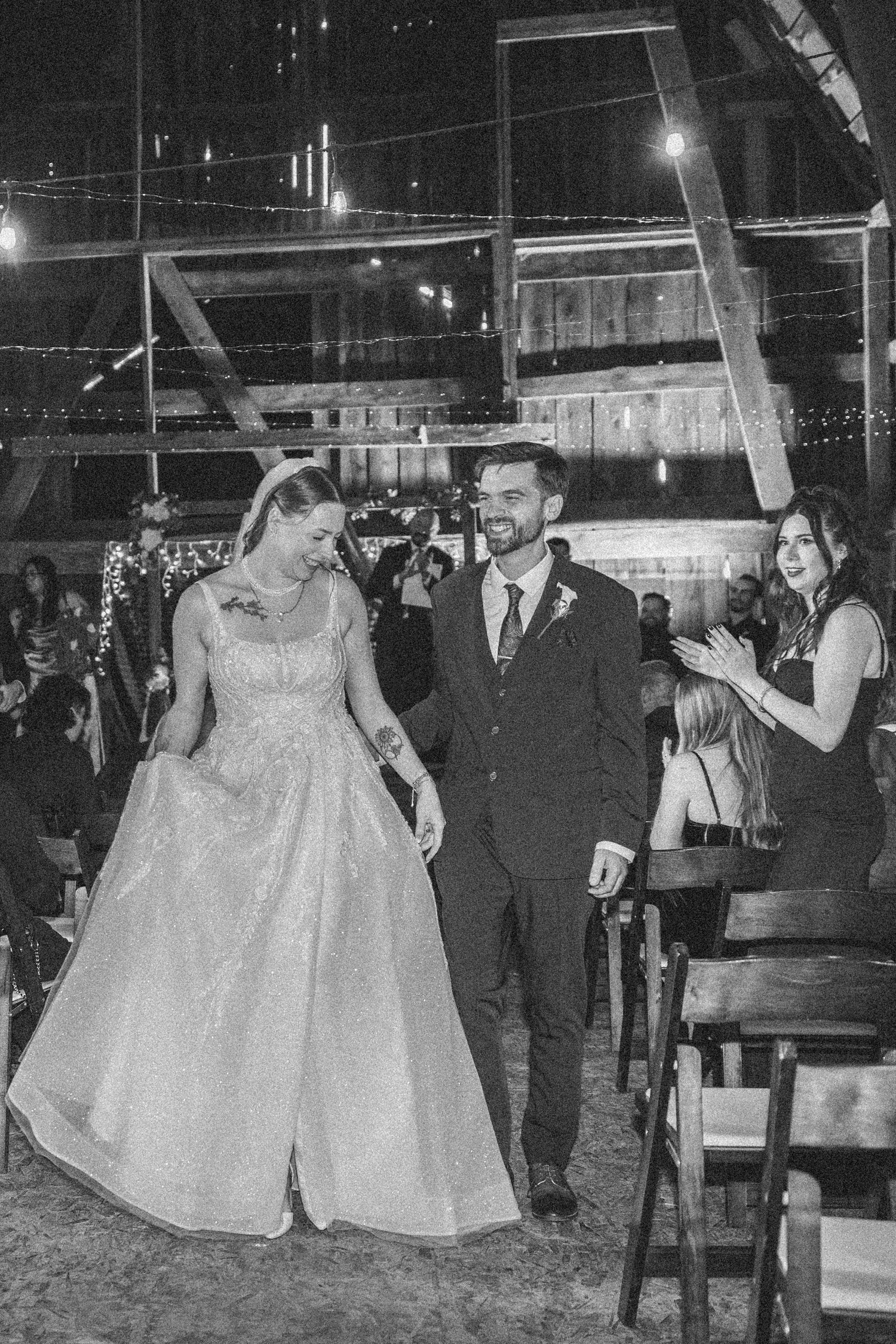 Newlyweds walk down the aisle at an indoor evening reception with string lights and rustic decor.
