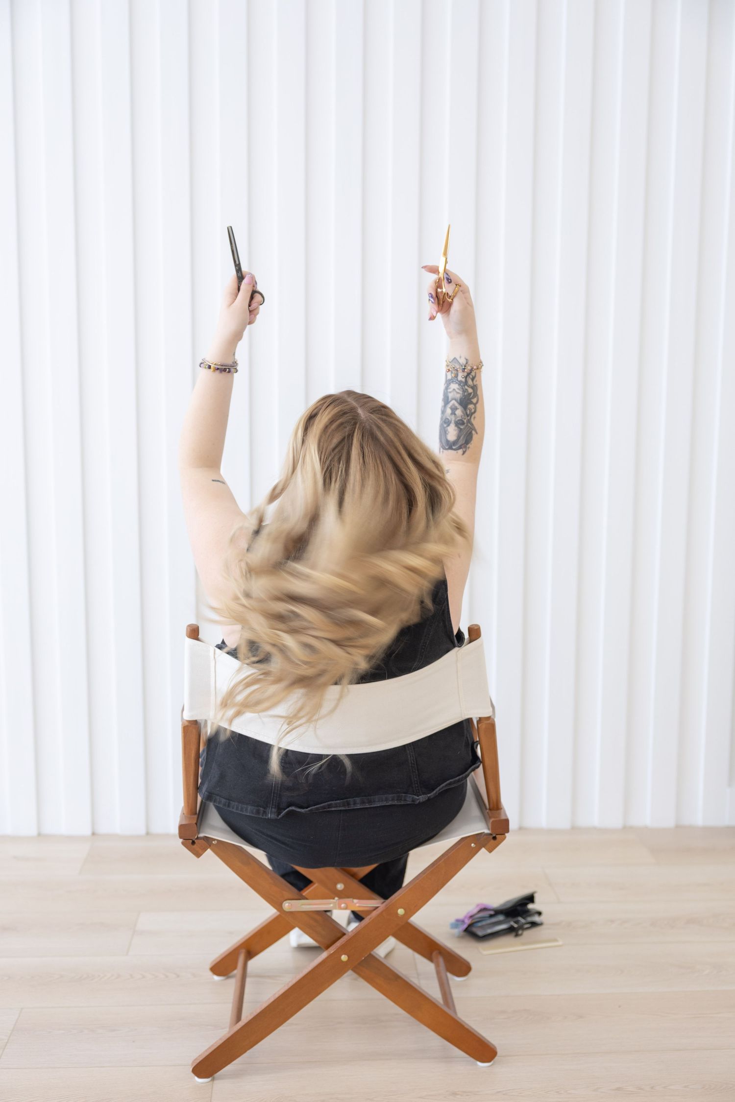 Blonde woman with tattoos sits in a wooden director's chair, arms raised holding a pencil and phone, viewed from behind.