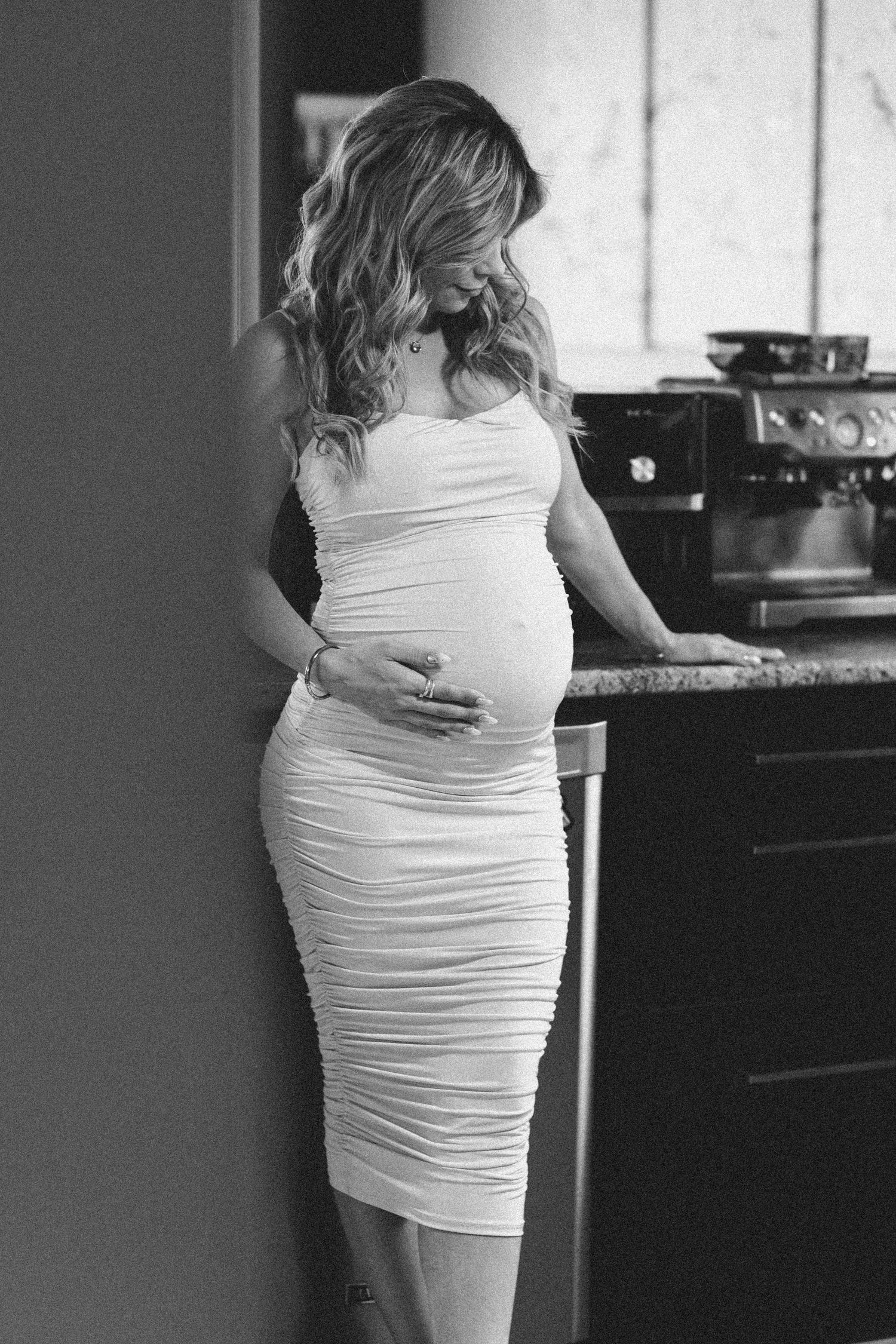 Black and white photo of a pregnant woman in a white ruched dress cradling her baby bump in a kitchen.