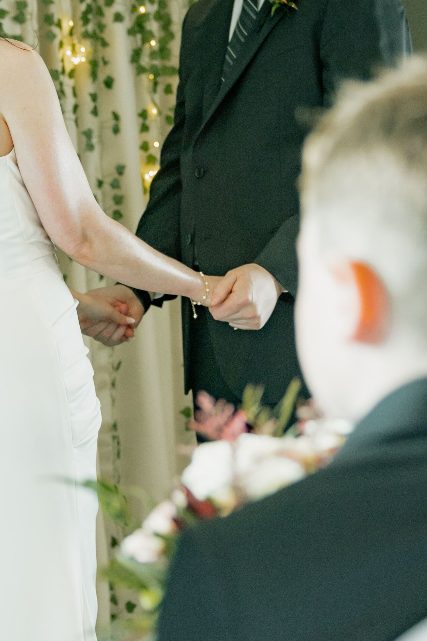 Bride and groom holding hands during wedding ceremony with ivy backdrop and fairy lights.