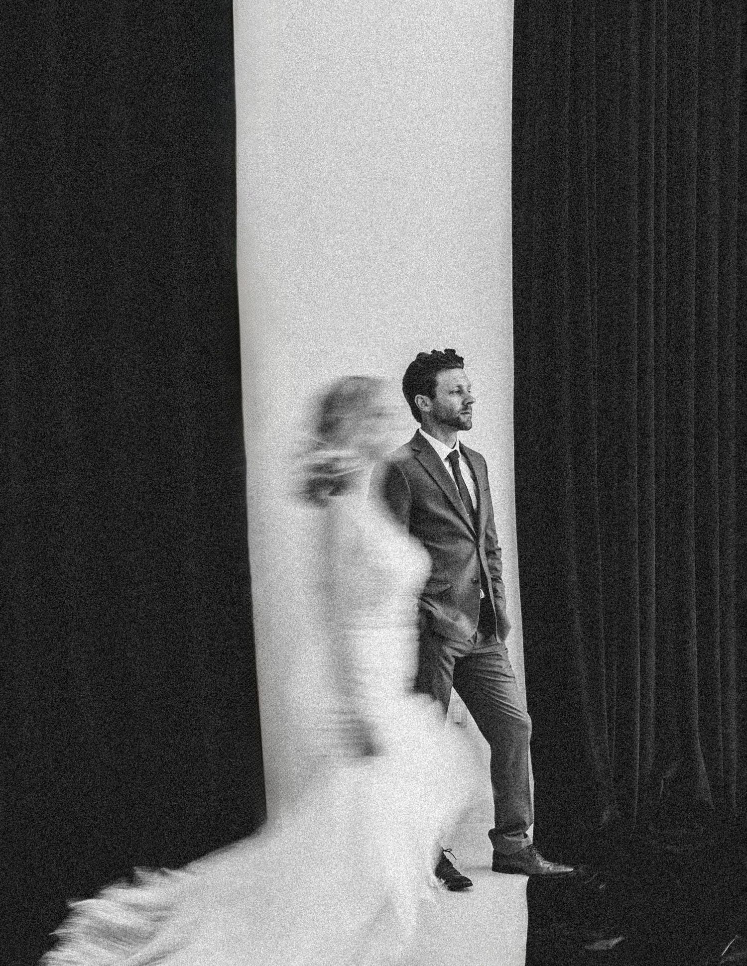 Artistic black and white photograph showing a figure standing by a pillar with a blurred motion effect in the foreground.