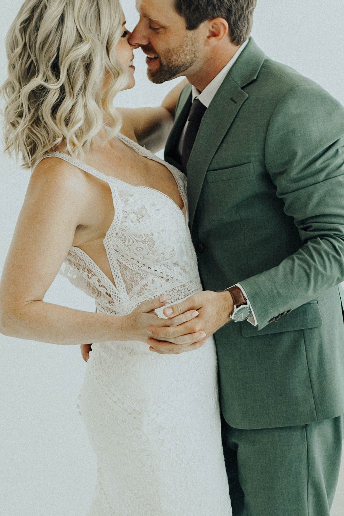 Romantic wedding couple embraces in intimate moment, with bride in lace gown and groom wearing green suit.