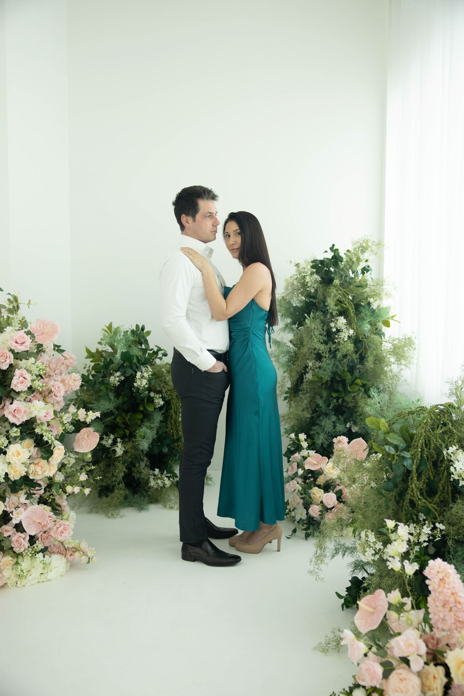 A couple in formal attire shares an intimate moment in a romantic studio setting with pink roses and greenery.