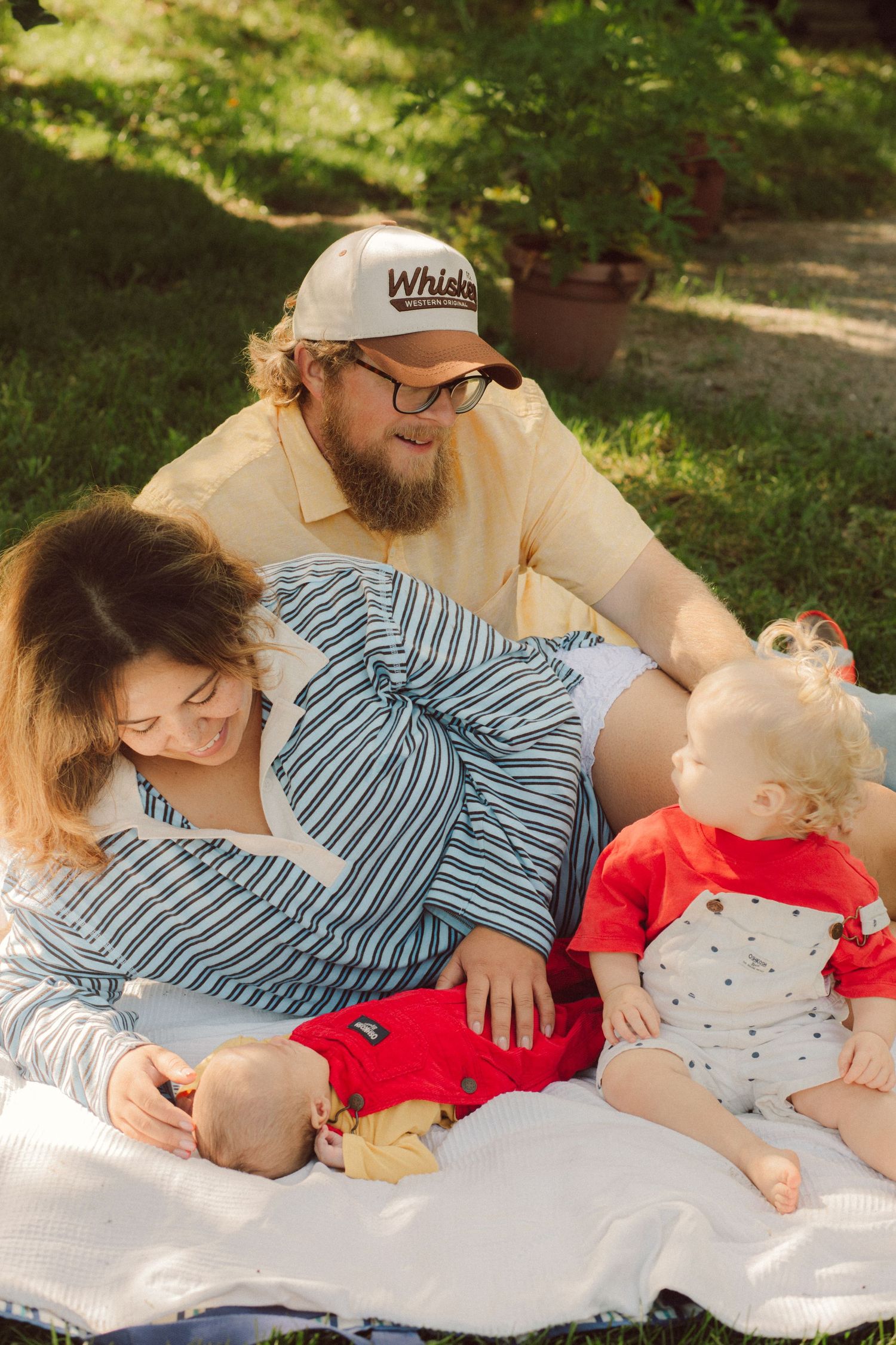 Family with two young children enjoying a sunny outdoor picnic on a blanket in a green garden.
