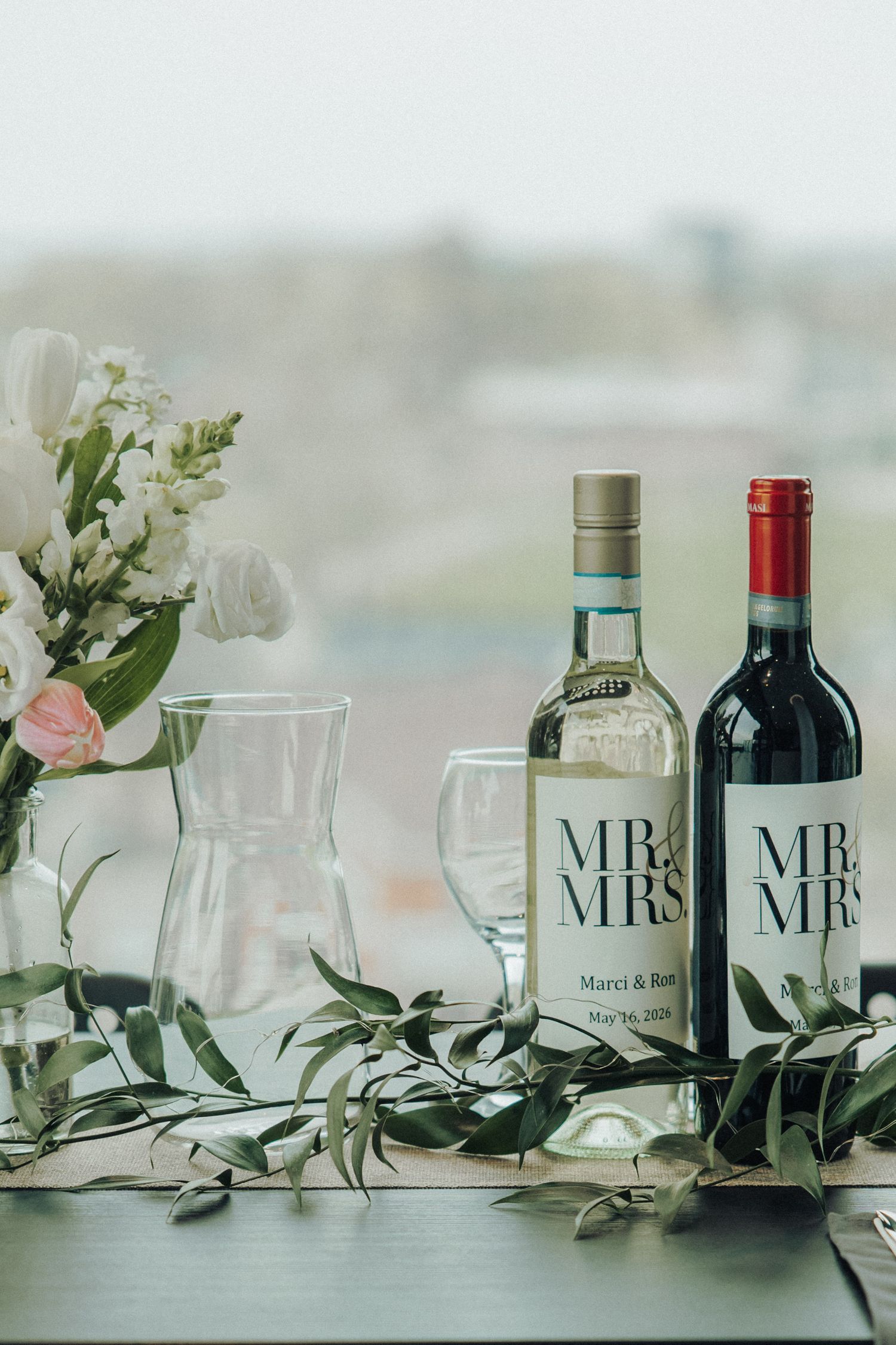 Two custom Mr. & Mrs. wedding wine bottles displayed with white flowers and greenery on a table.