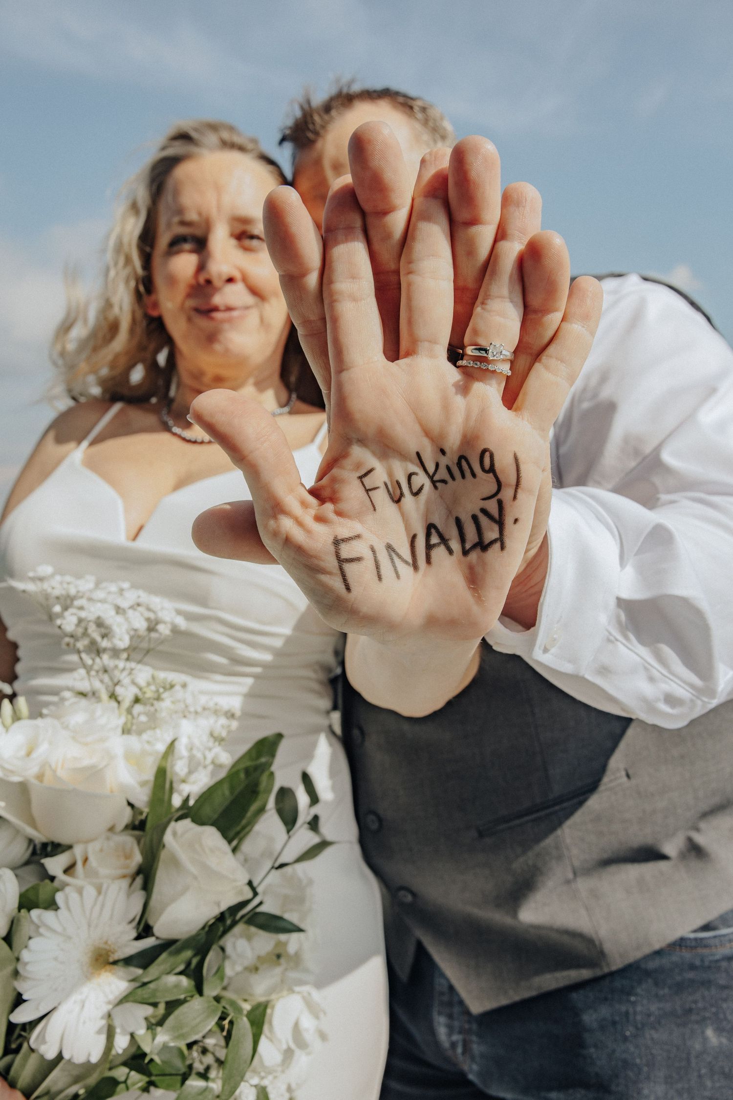 Newlyweds showing palm with Fucking Finally written on it, celebrating their wedding day outdoors.