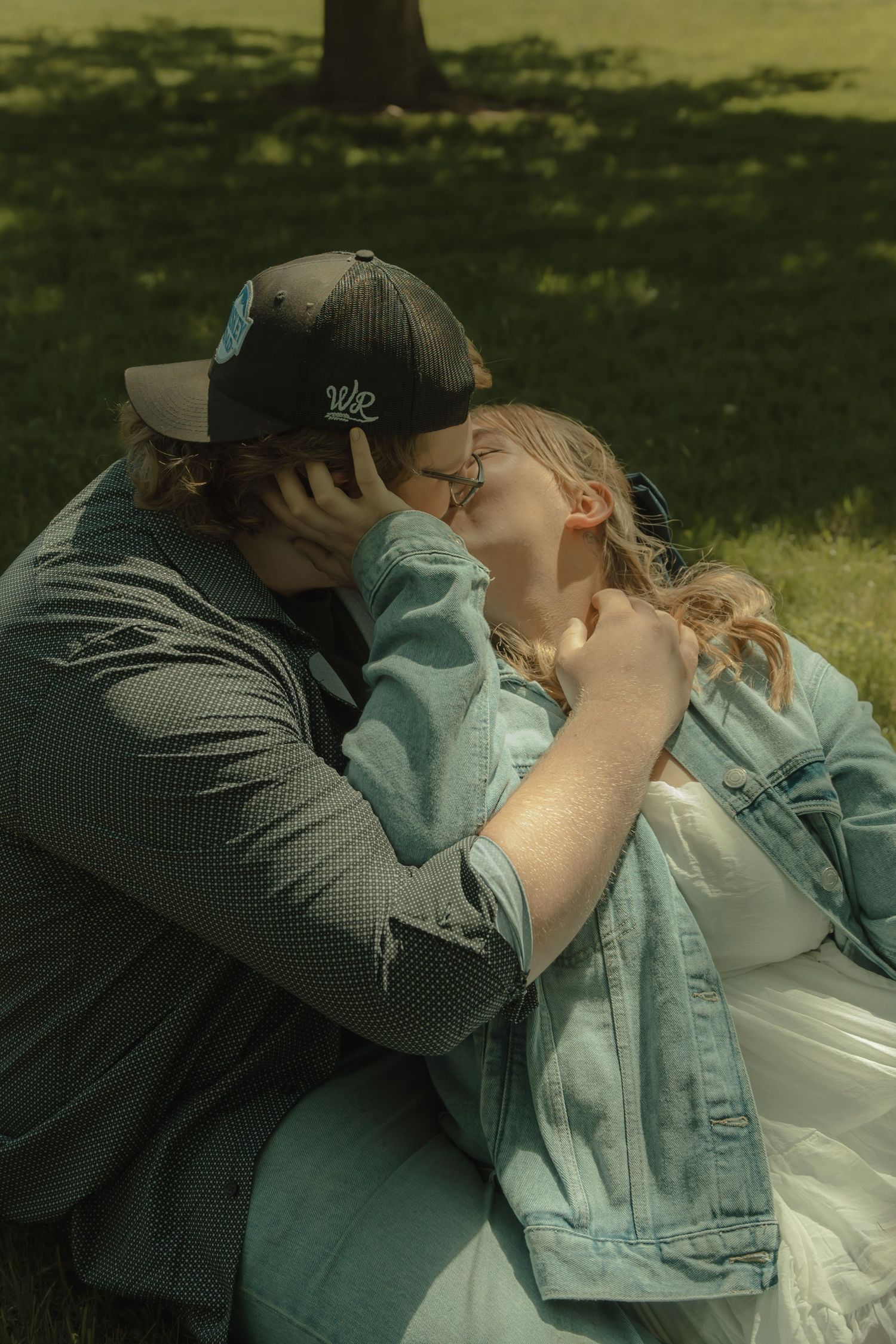 Couple sharing a tender kiss while sitting on green grass in a sunny park, woman in denim jacket and man in snapback cap.