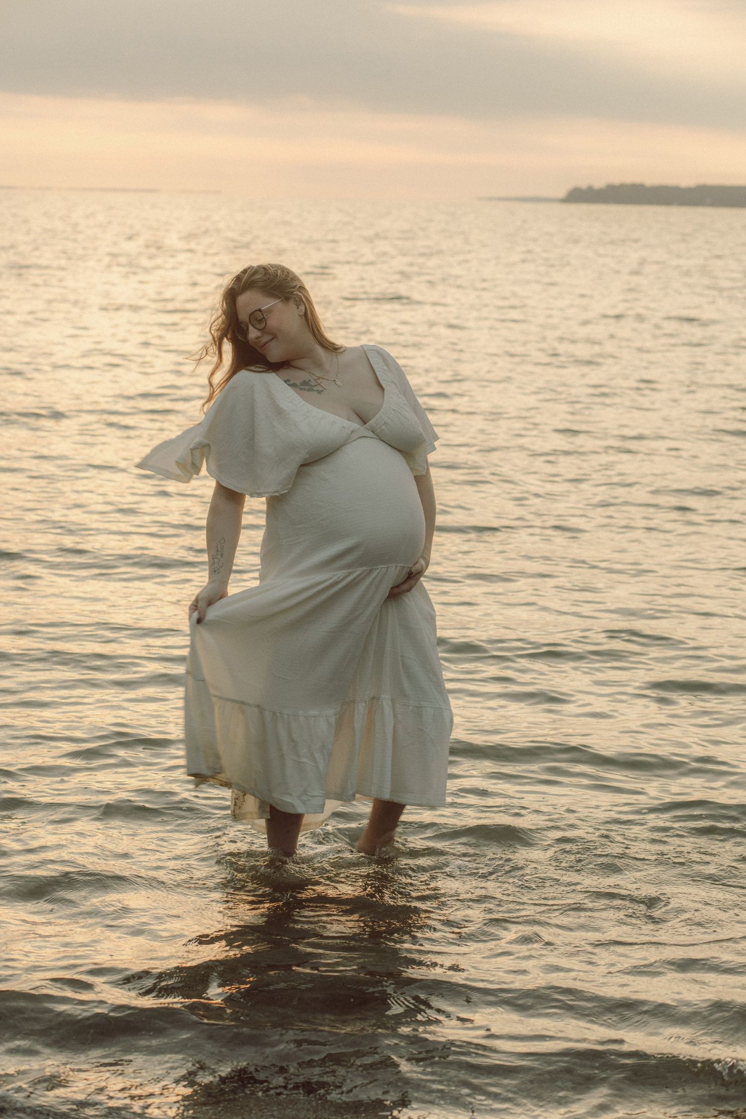Pregnant woman in white dress standing in calm ocean water at golden sunset, cradling her baby bump with a serene smile.