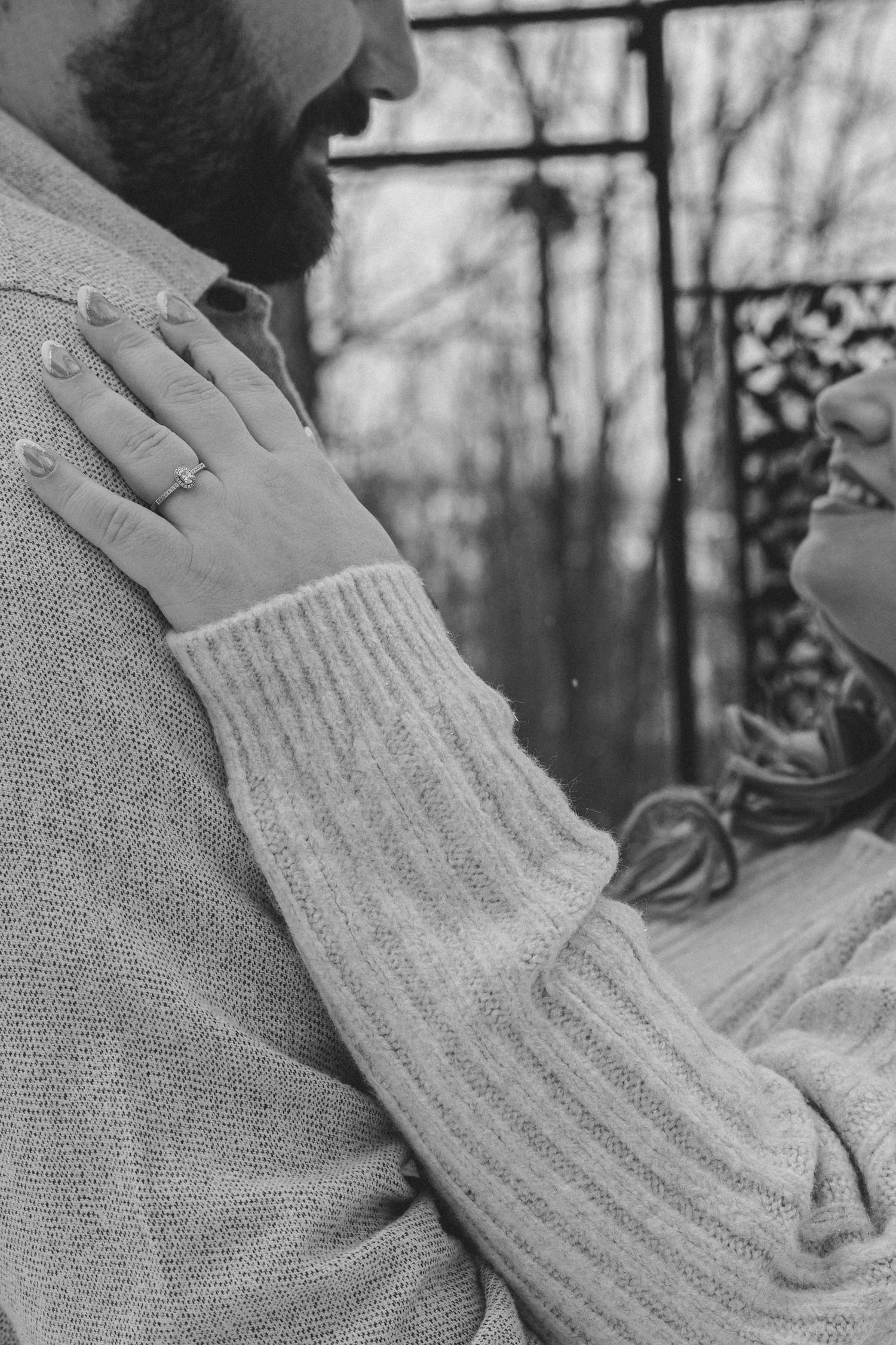 A close-up black and white photo showing hands resting on a sweater-clad shoulder against a blurred outdoor background.