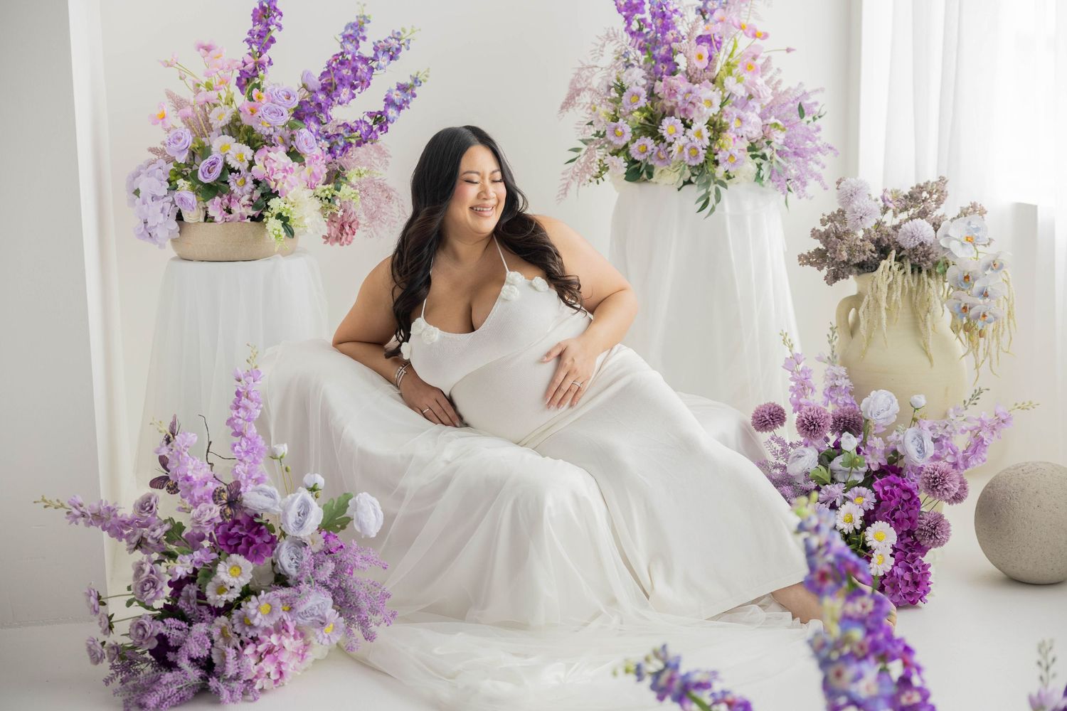 A romantic bridal photoshoot featuring flowing white dress and purple floral arrangements in a bright studio setting.