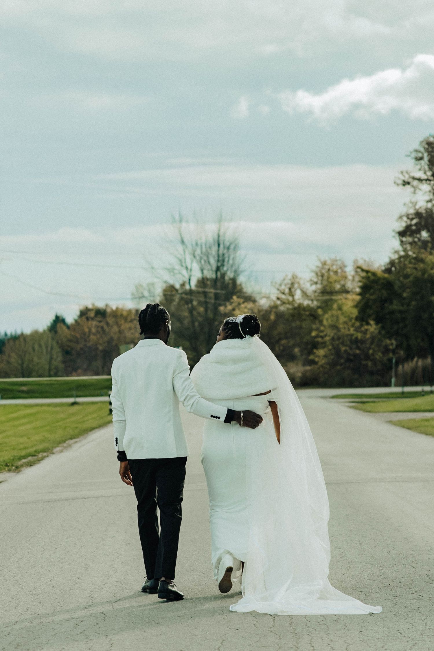 A couple in wedding attire walking away on a paved path surrounded by autumn trees and grass.