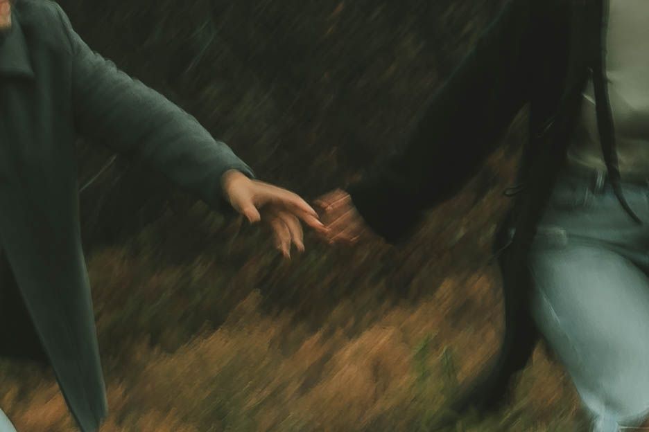 A person in a green jacket and jeans walks through an autumn forest with fallen leaves on the ground.