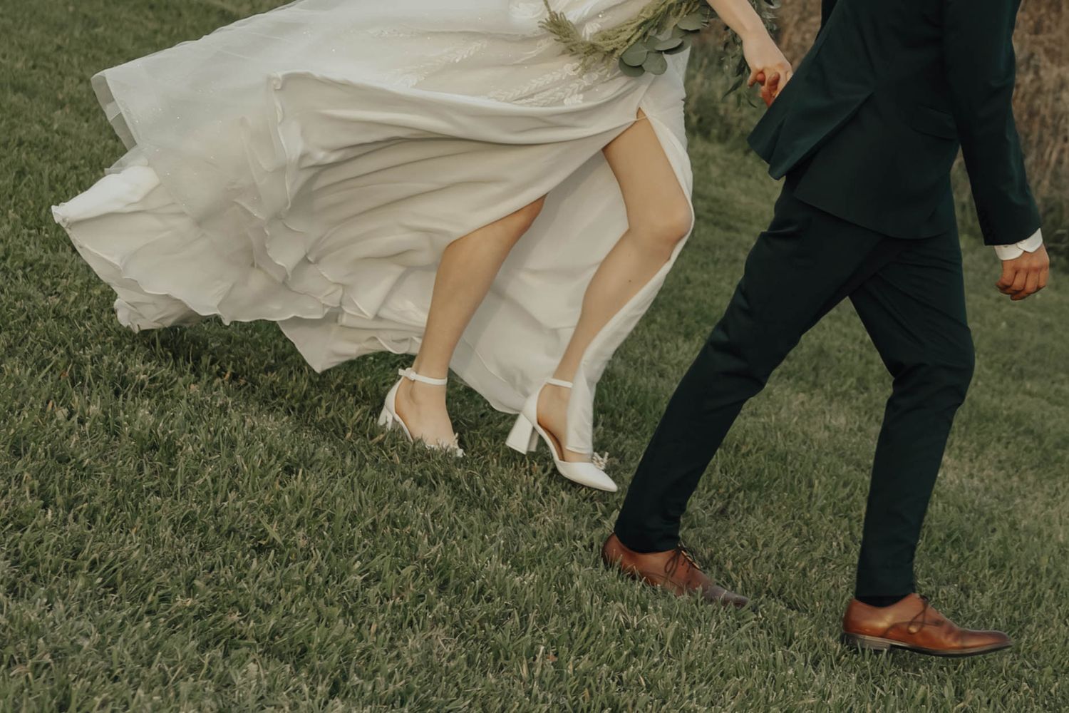 A bride's flowing white dress and groom's dress shoes captured in motion while walking on grass at a wedding.