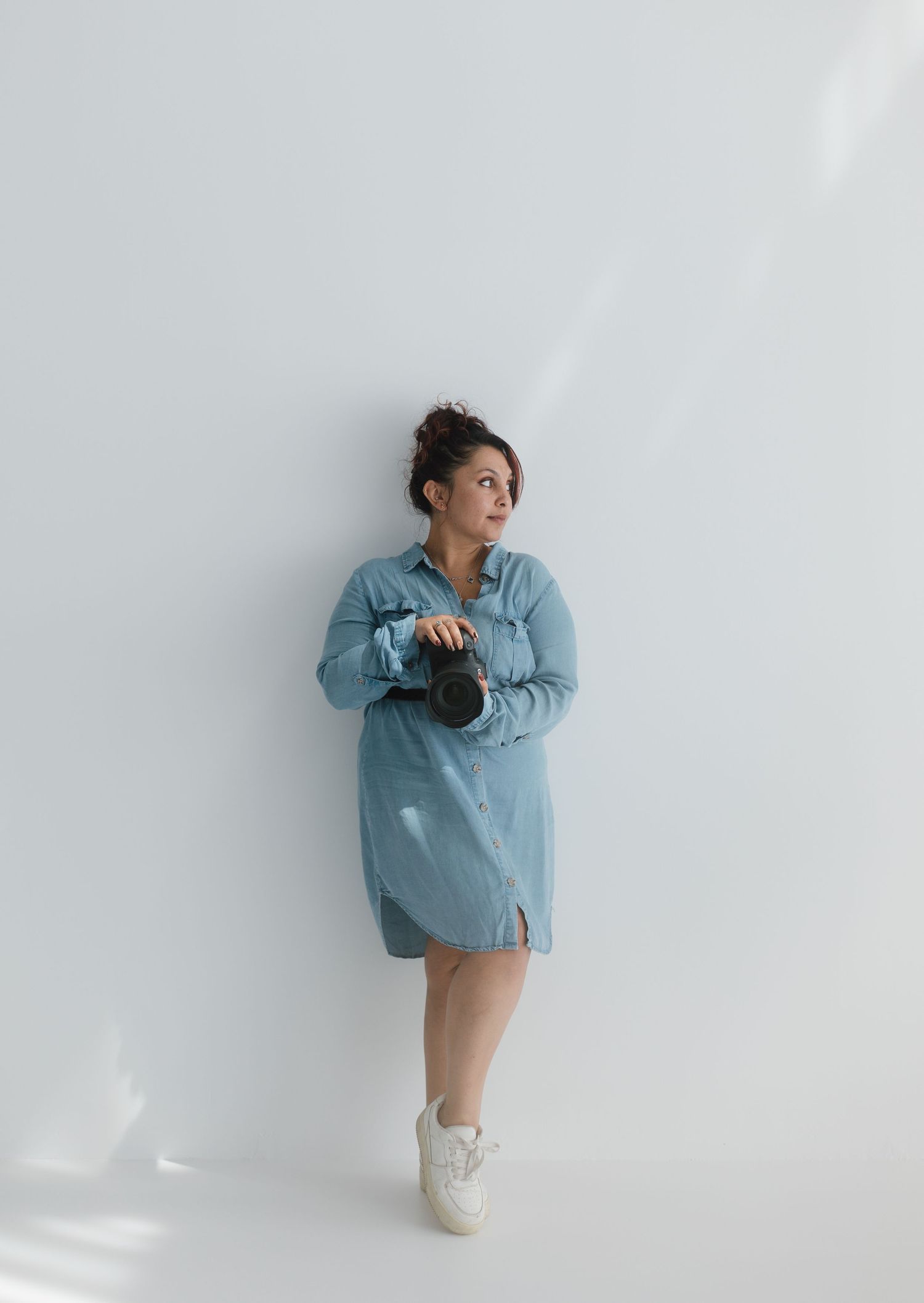 Woman in blue denim shirt dress holding a camera, leaning against a white wall wearing white sneakers.