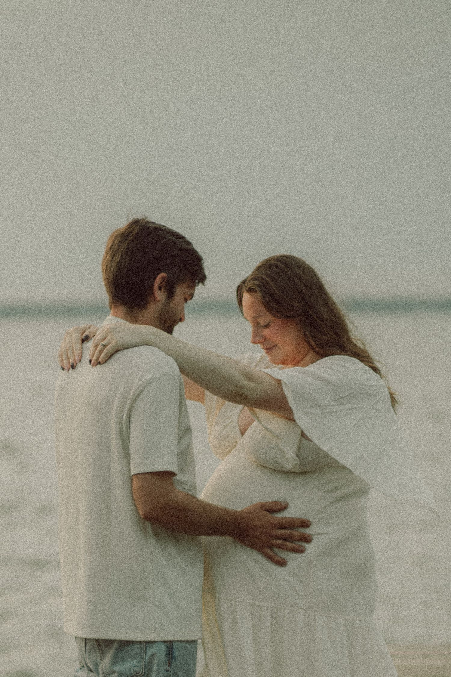 Pregnant couple embracing on beach, woman in white dress with hands on partner's shoulders near ocean.