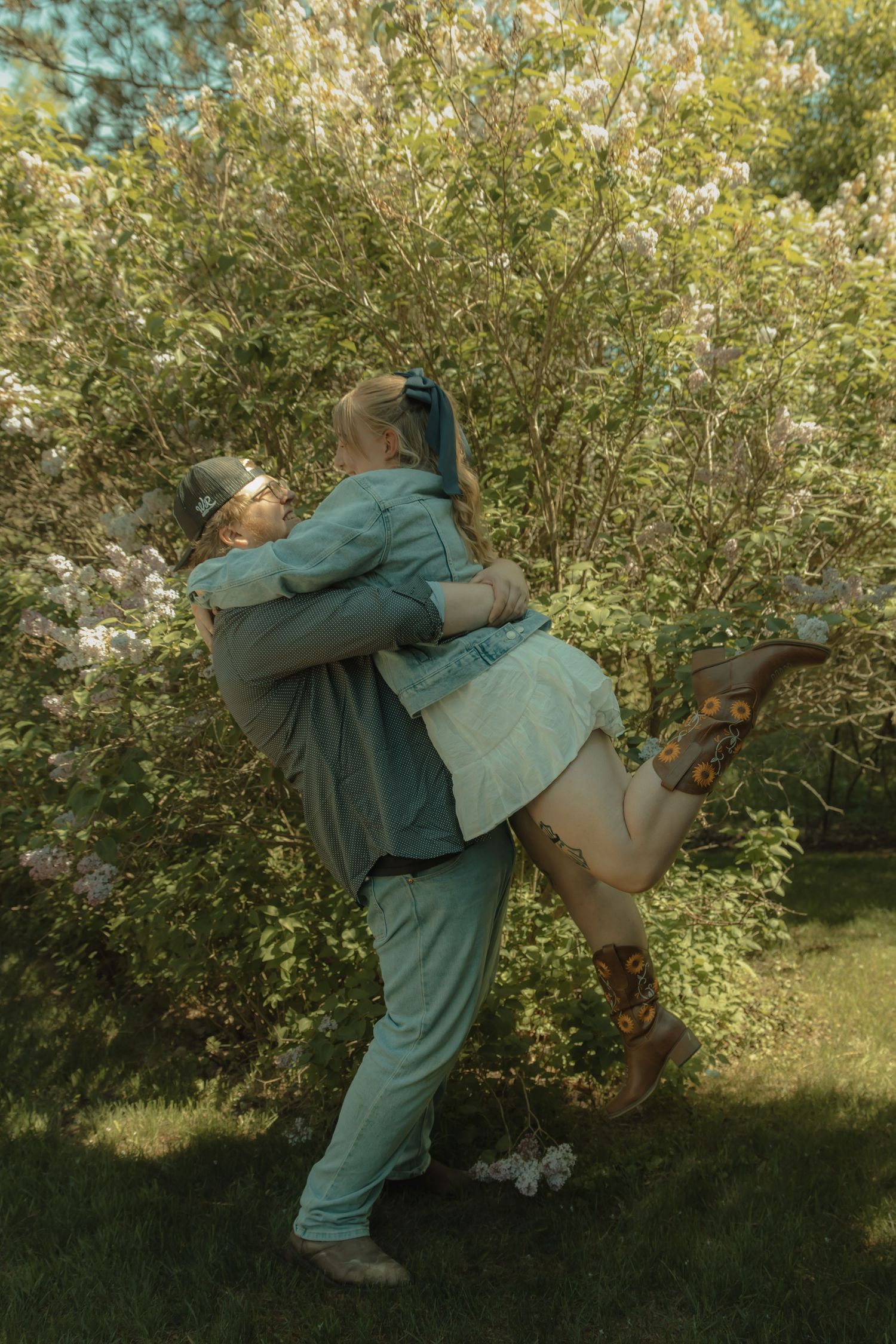 A couple embraces in a romantic lift pose among blooming lilac bushes in a lush green garden.