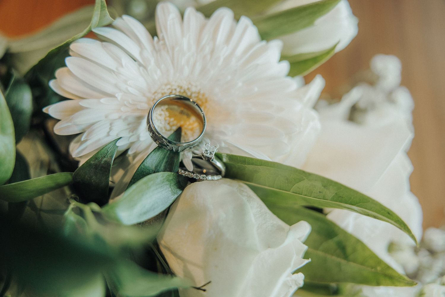 Two wedding rings resting on a white gerbera daisy bouquet with green leaves and roses.