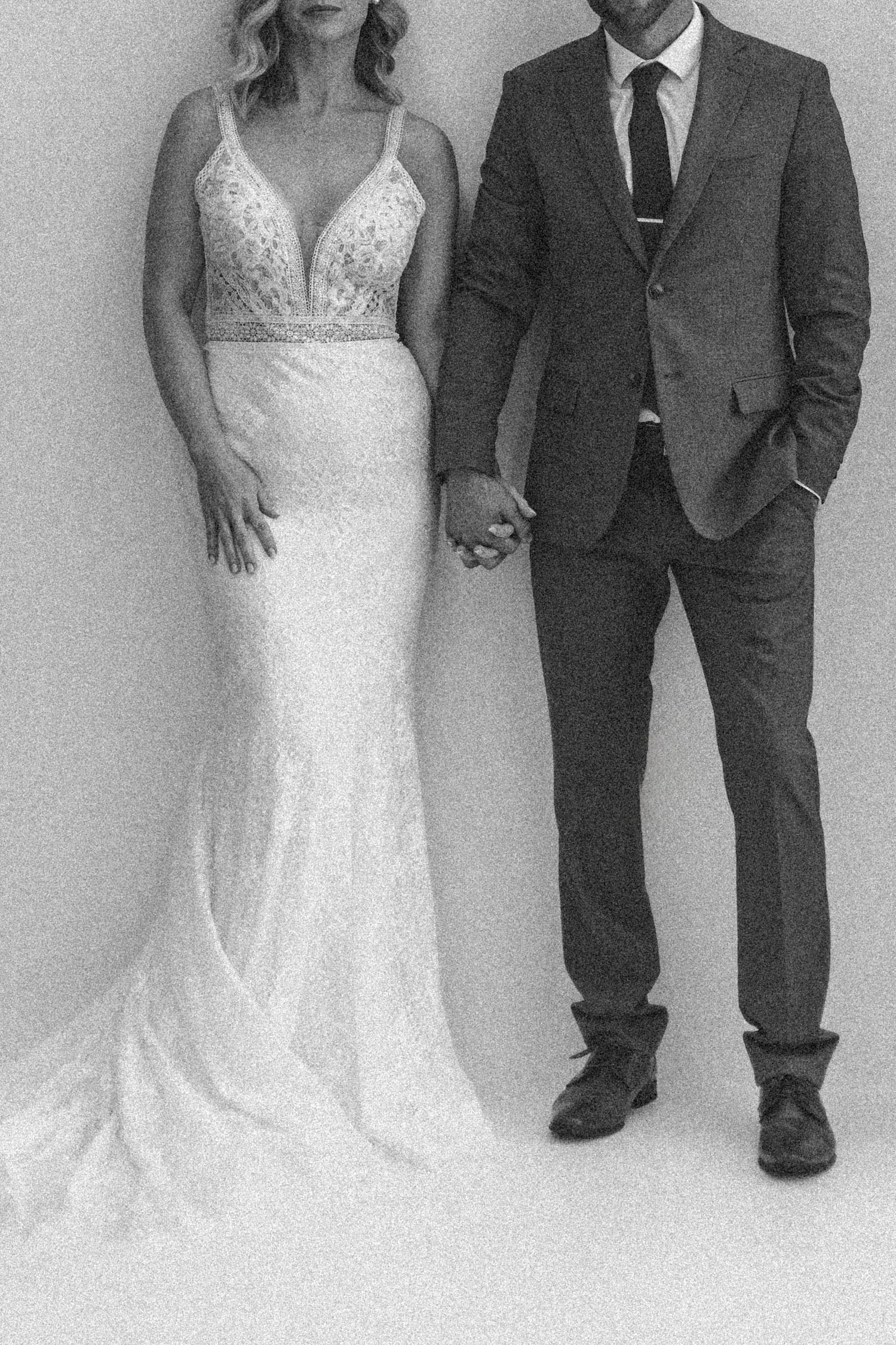 A black and white wedding photo showing a bride in a fitted lace gown holding hands with a groom in a dark suit.