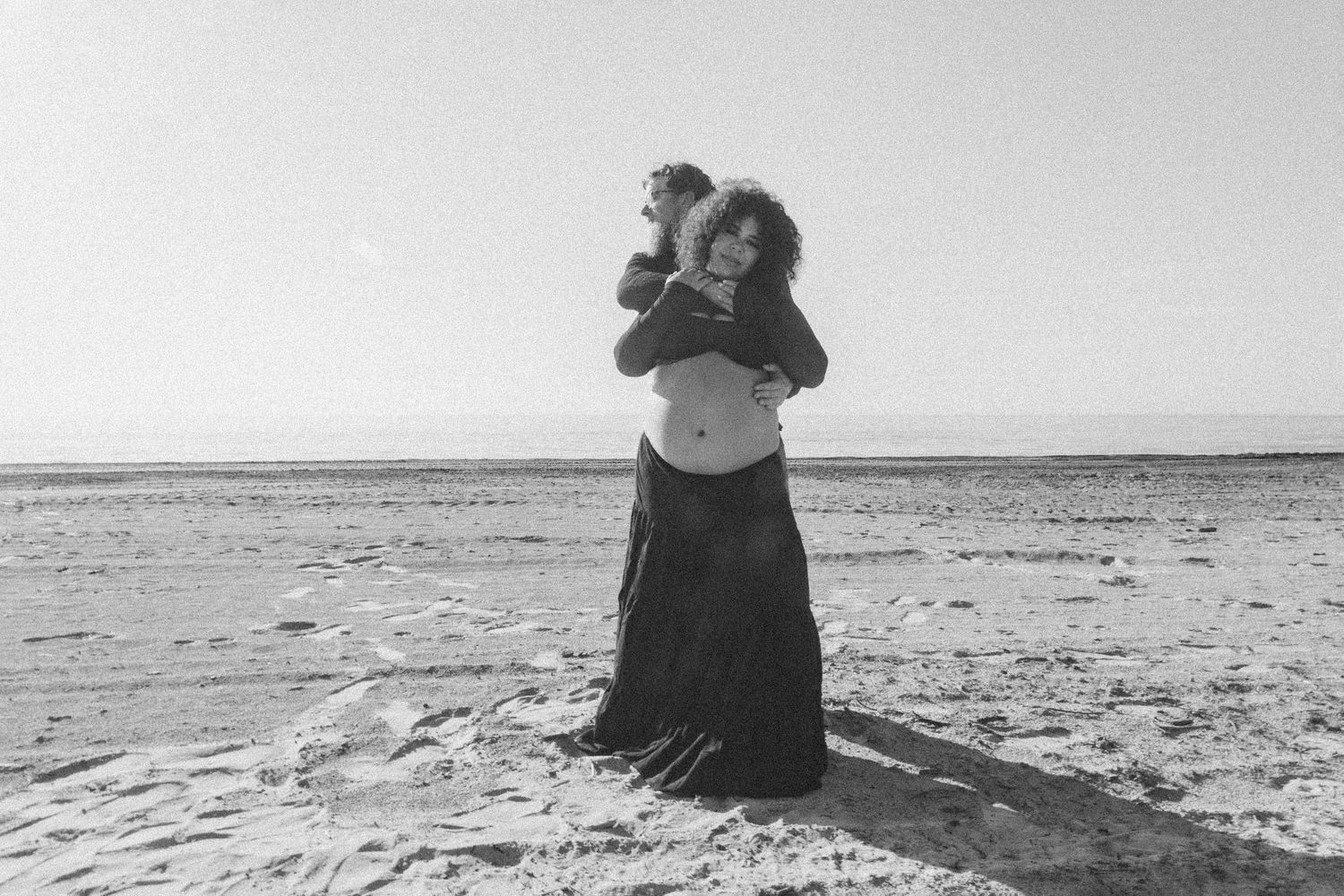 Black and white photo of a person in a long dress standing on a beach shore gazing through binoculars.