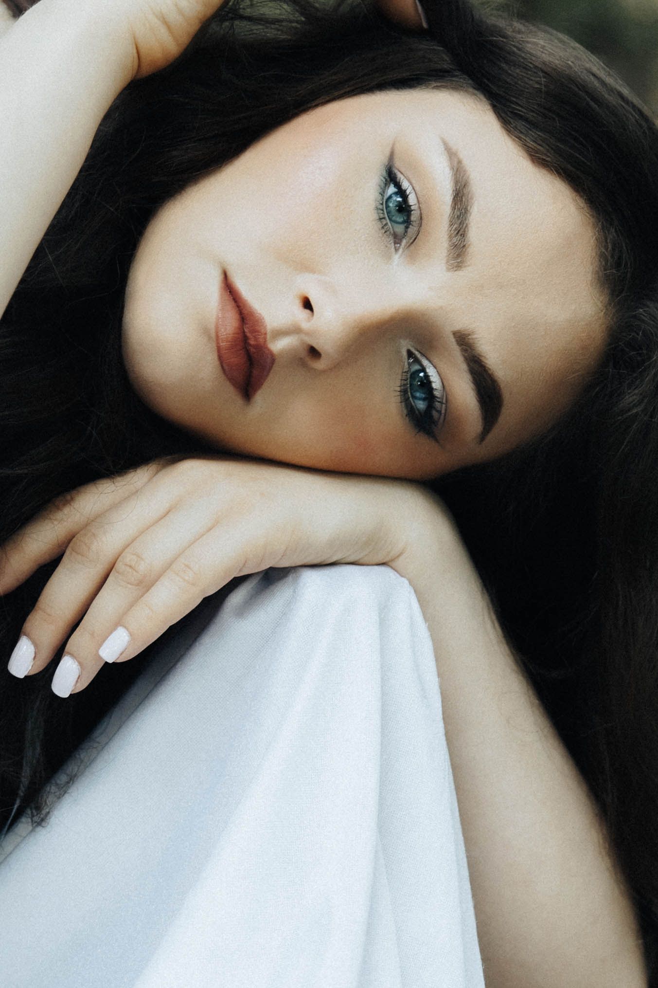 Close up portrait showing striking blue eyes, dark hair and red lips against white fabric.