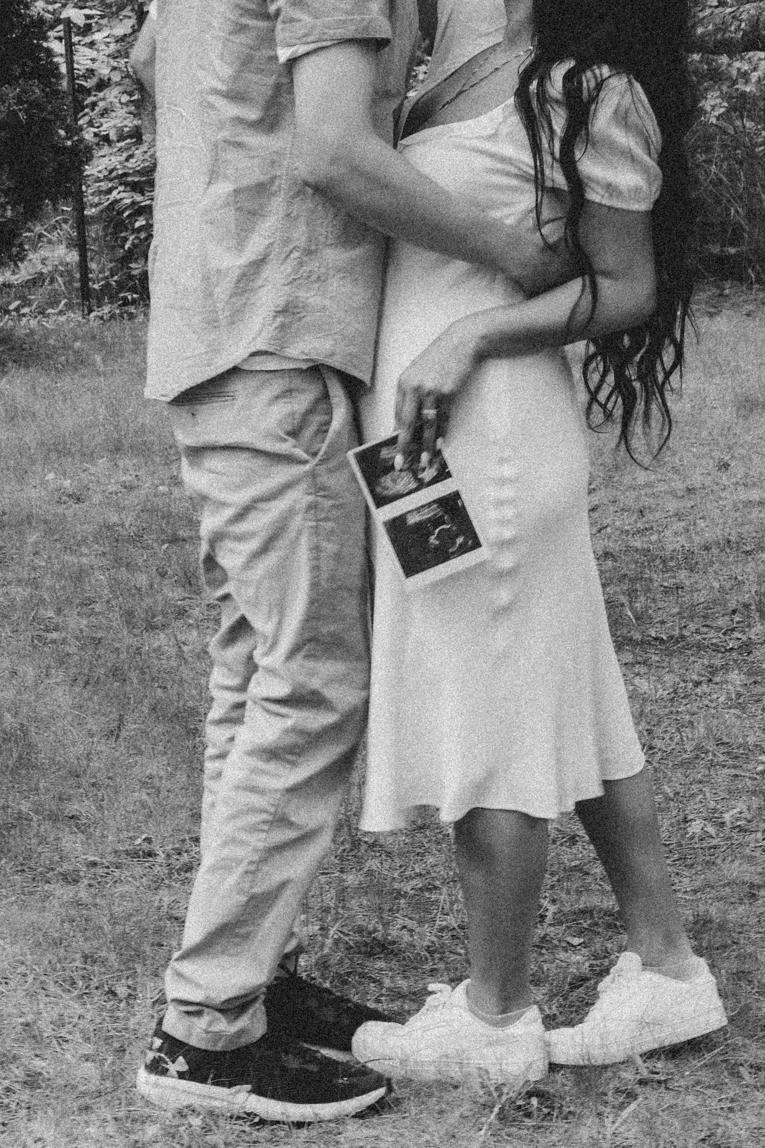 Black and white photo showing legs and hands holding a polaroid camera while embracing outdoors.