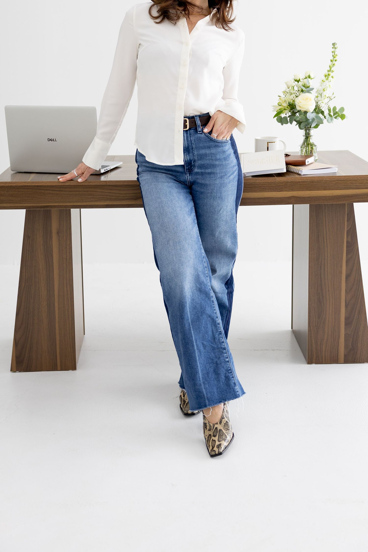 Woman in white button-up shirt, blue wide-leg jeans, and snakeskin boots leaning against a wooden desk.