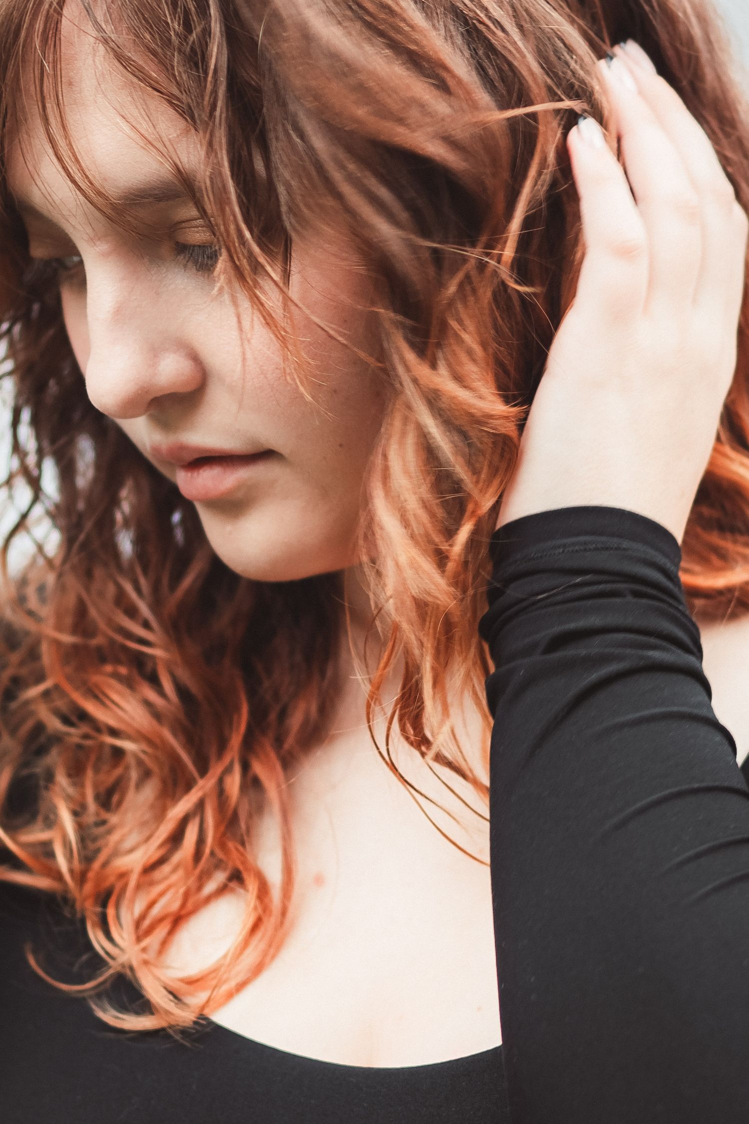 Young woman with red wavy hair wearing a black top, eyes closed, hand raised to her hair.