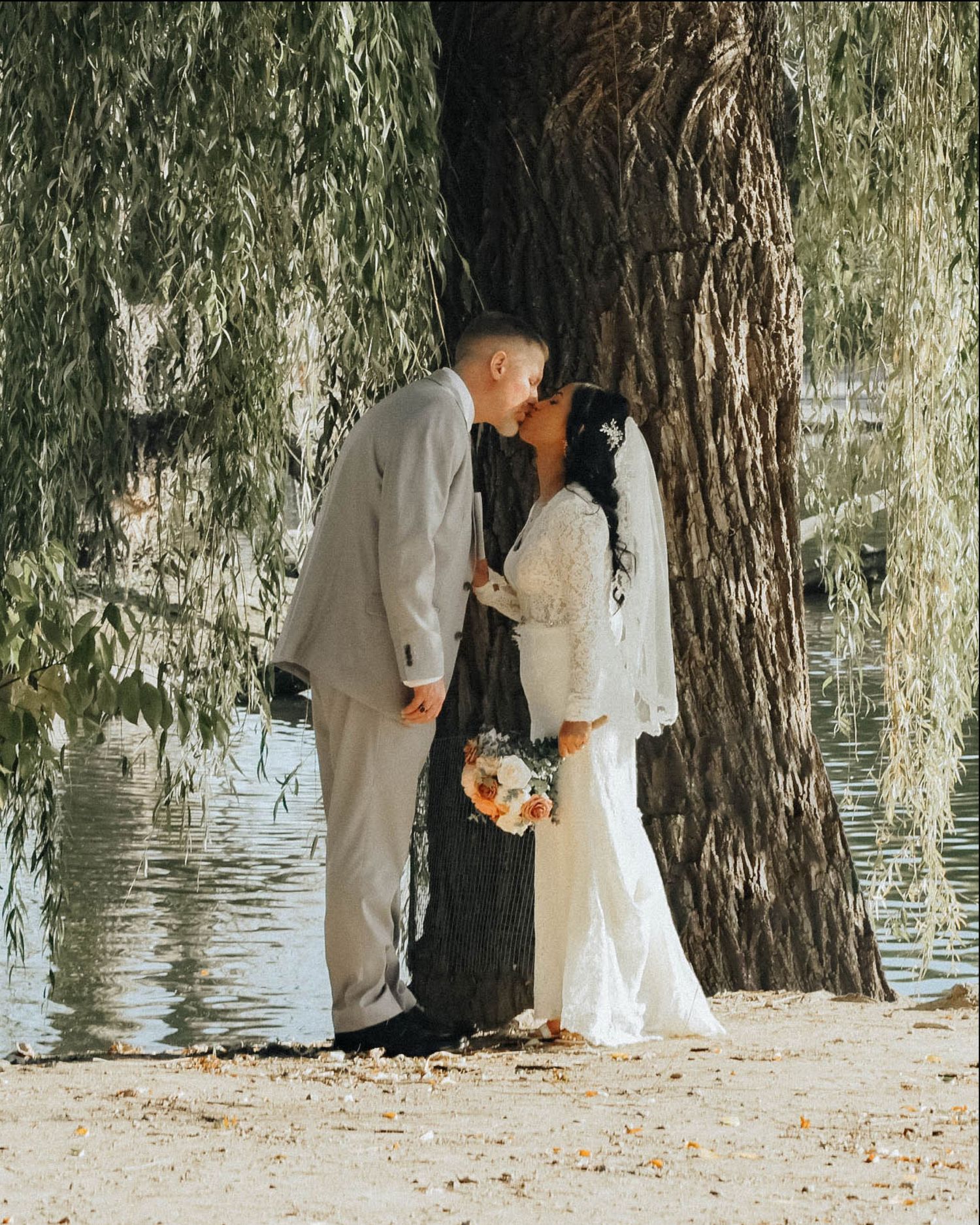 A romantic wedding couple embraces under a weeping willow tree beside a tranquil pond.