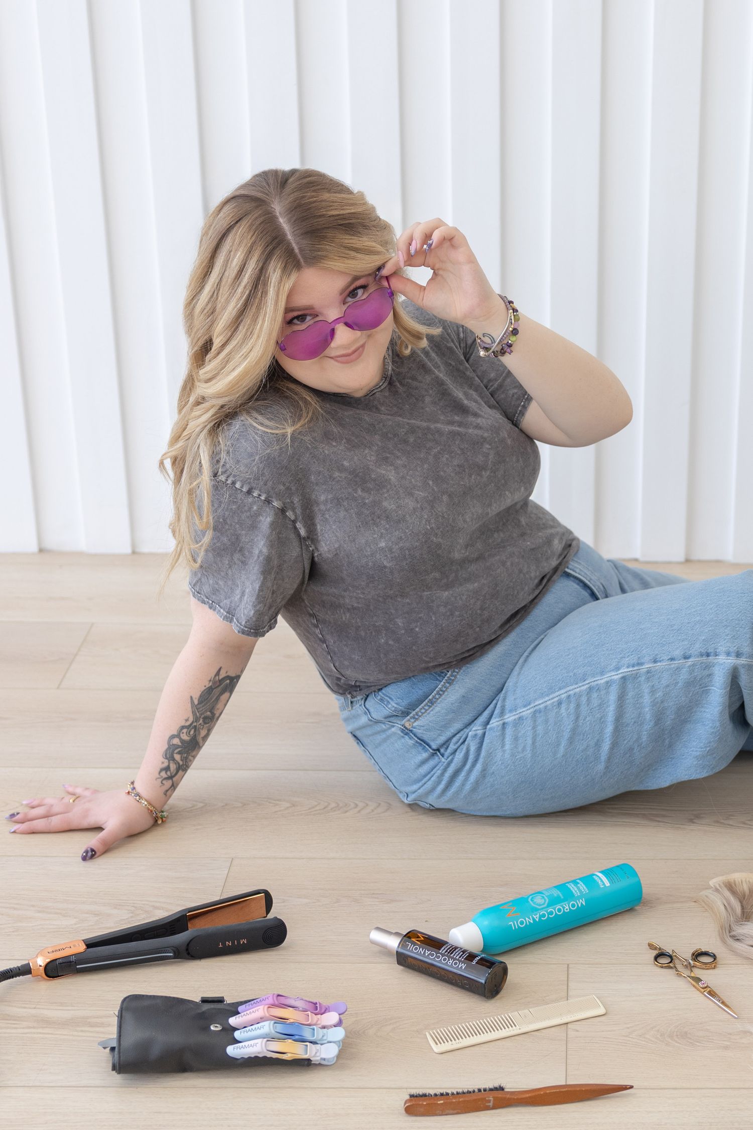 Woman with tattoos and pink sunglasses sitting on floor surrounded by hair styling tools and accessories.