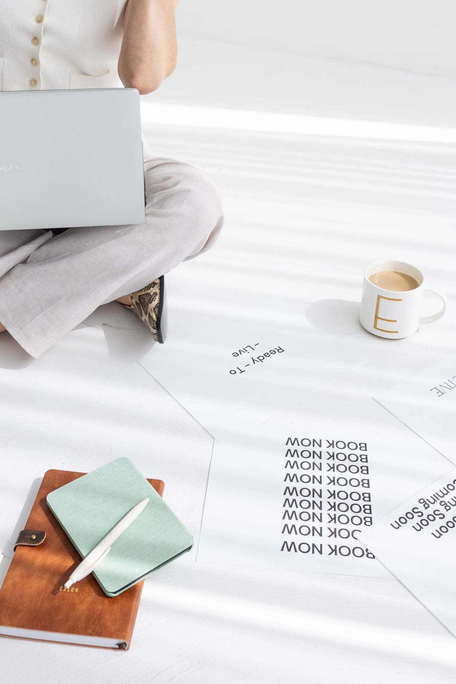 Person sitting cross-legged with laptop, coffee mug, notebooks, and printed booking pages on white floor.