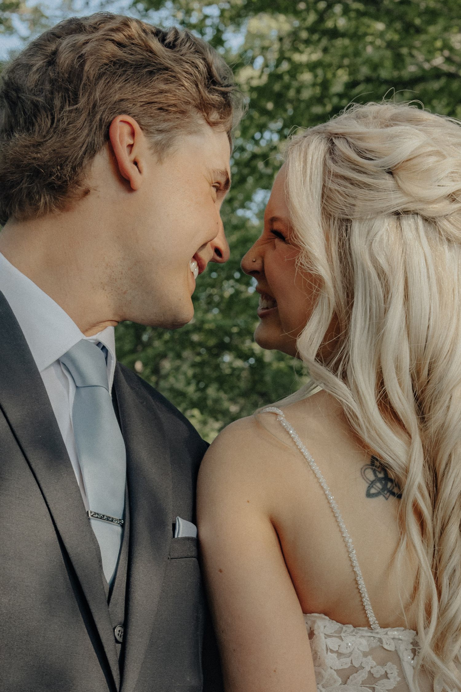 A smiling bride and groom in wedding attire share a romantic close moment outdoors with green trees in the background.