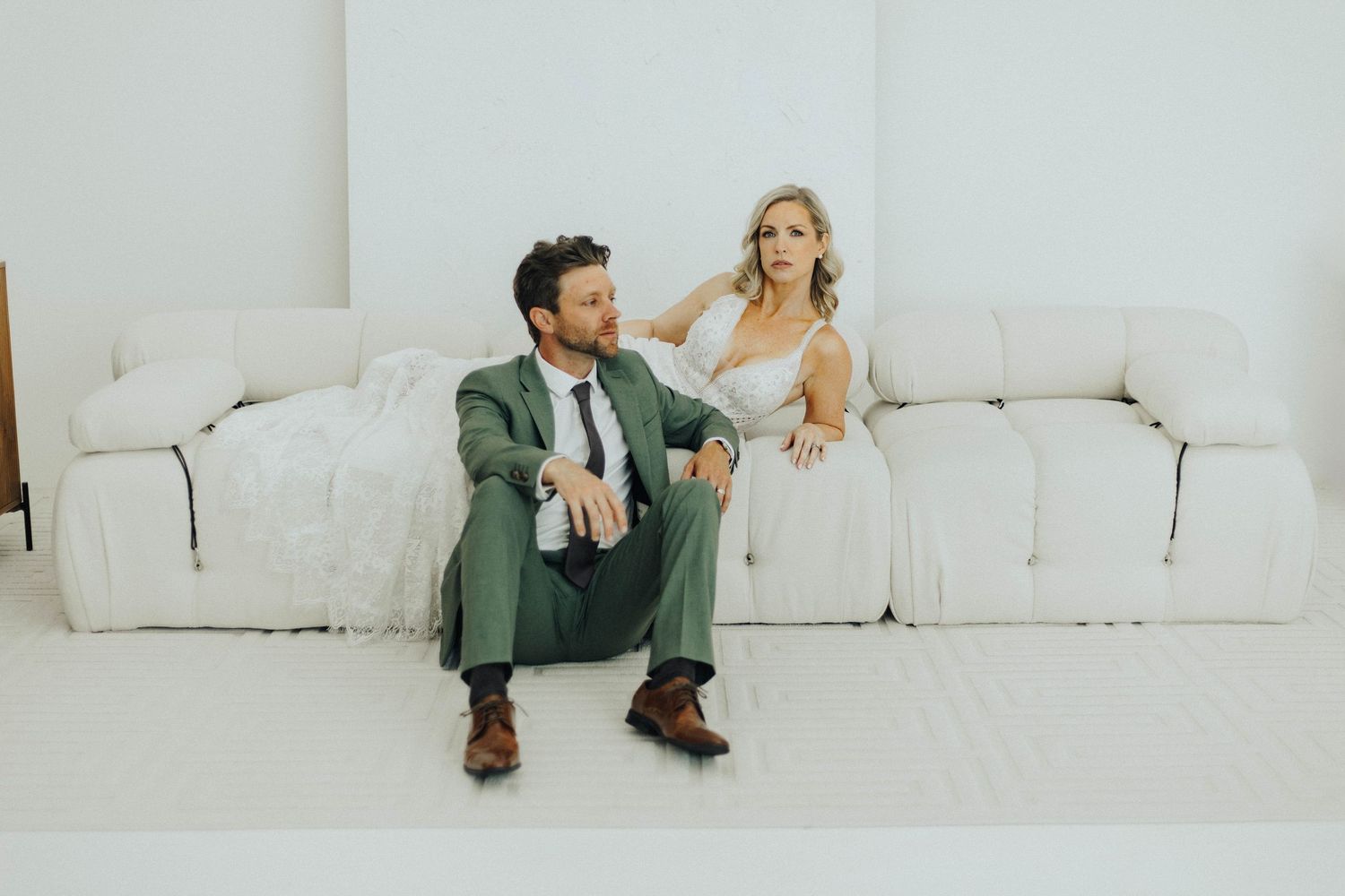 Couple in formal attire sits casually on a modern white couch against a minimalist white background.