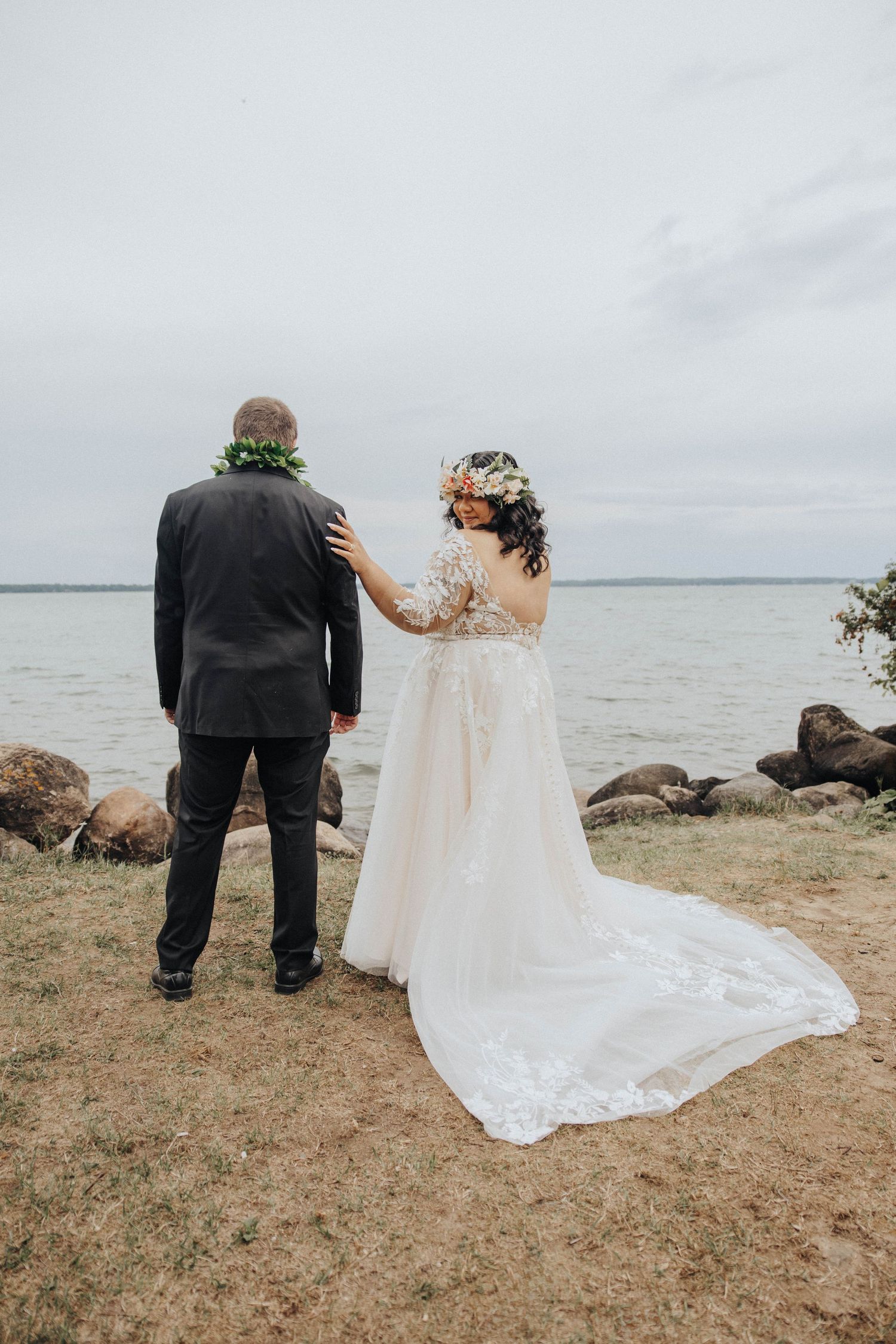 A wedding couple stands by a rocky shoreline with an overcast sky and calm waters in the background.