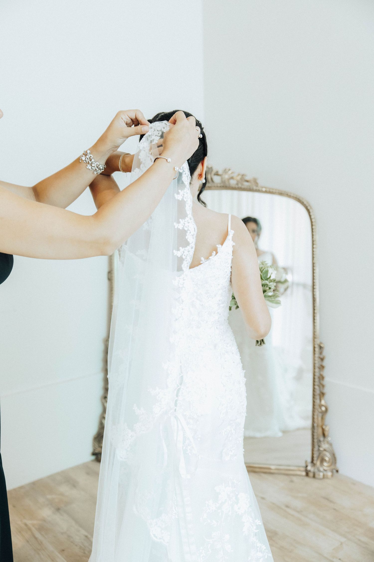 A close-up of hands adjusting a delicate white lace wedding dress in front of an ornate vintage mirror.