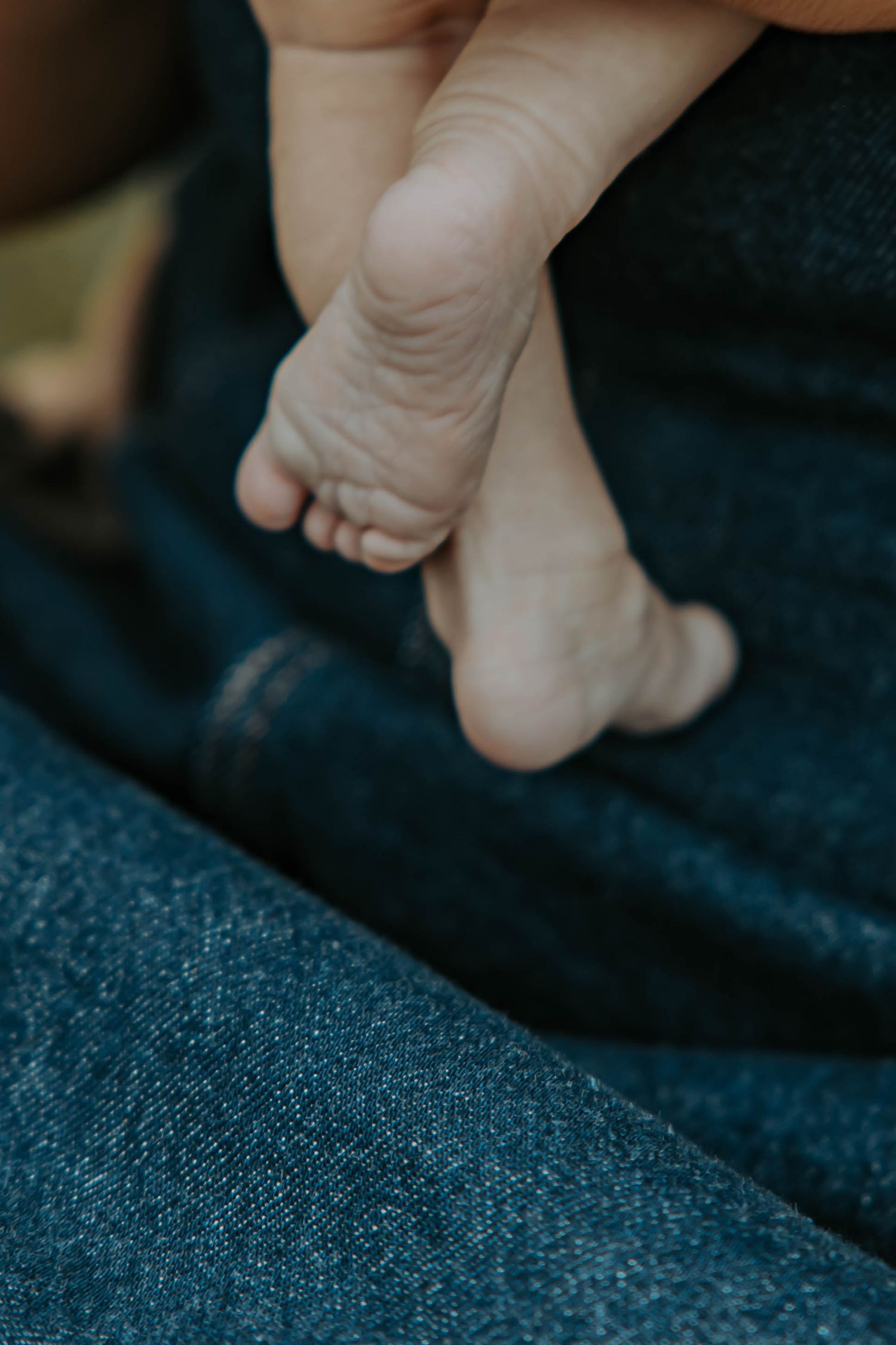 Close-up view of tiny baby feet resting on dark denim fabric showing delicate newborn toes and soft skin texture.