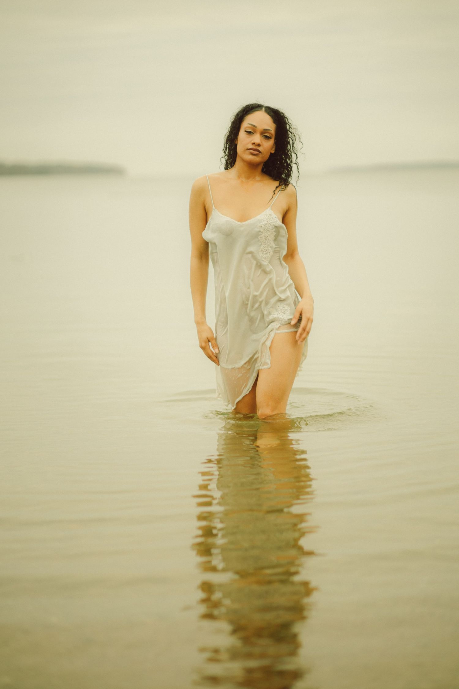 A woman in a wet white slip dress stands in shallow water, her reflection visible on the calm surface.