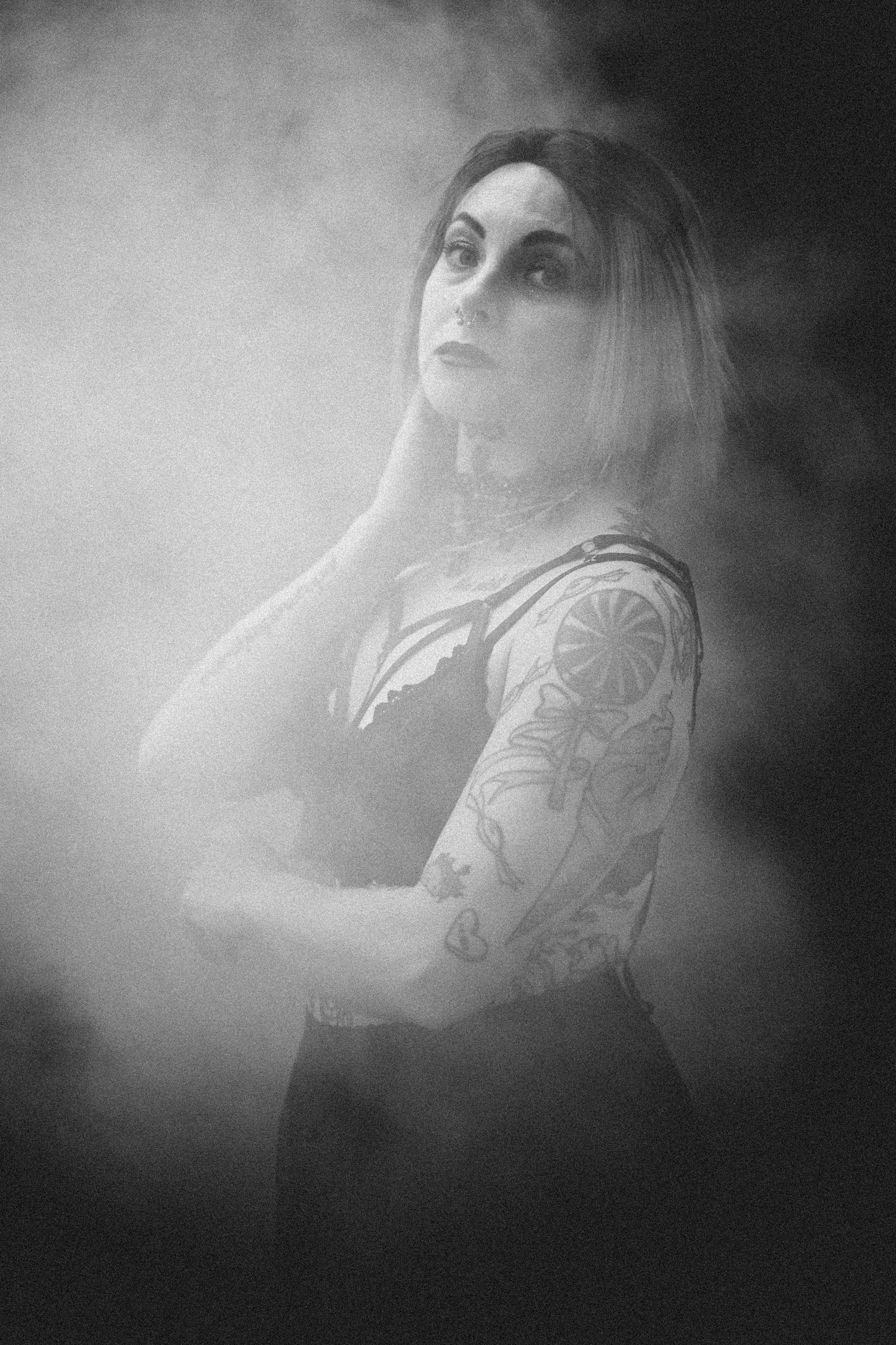 Black and white photo of a tattooed woman with long hair, arms crossed, surrounded by dramatic smoke.