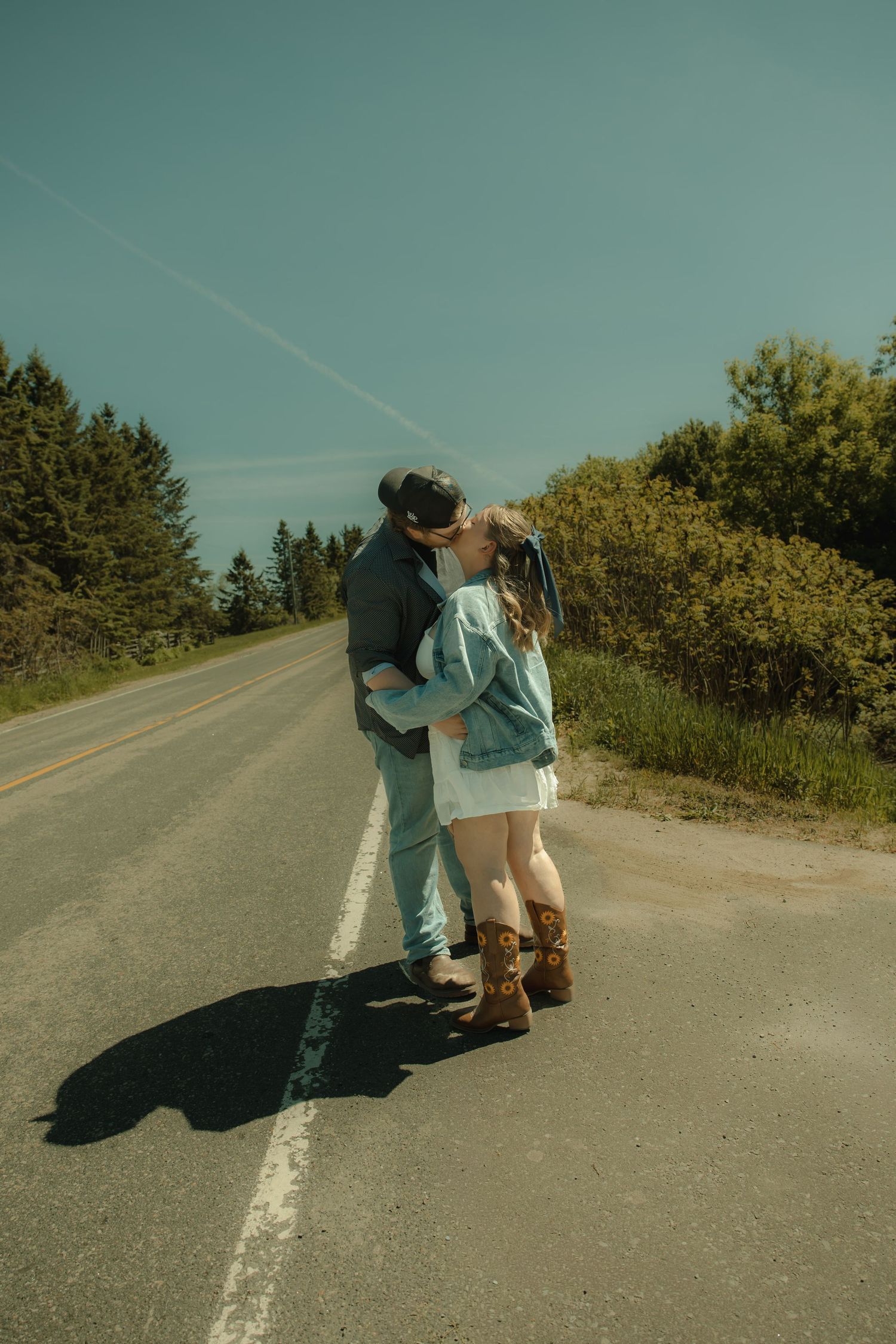 Romantic couple kissing on an empty road surrounded by trees, woman in denim jacket and brown boots, sunny day.
