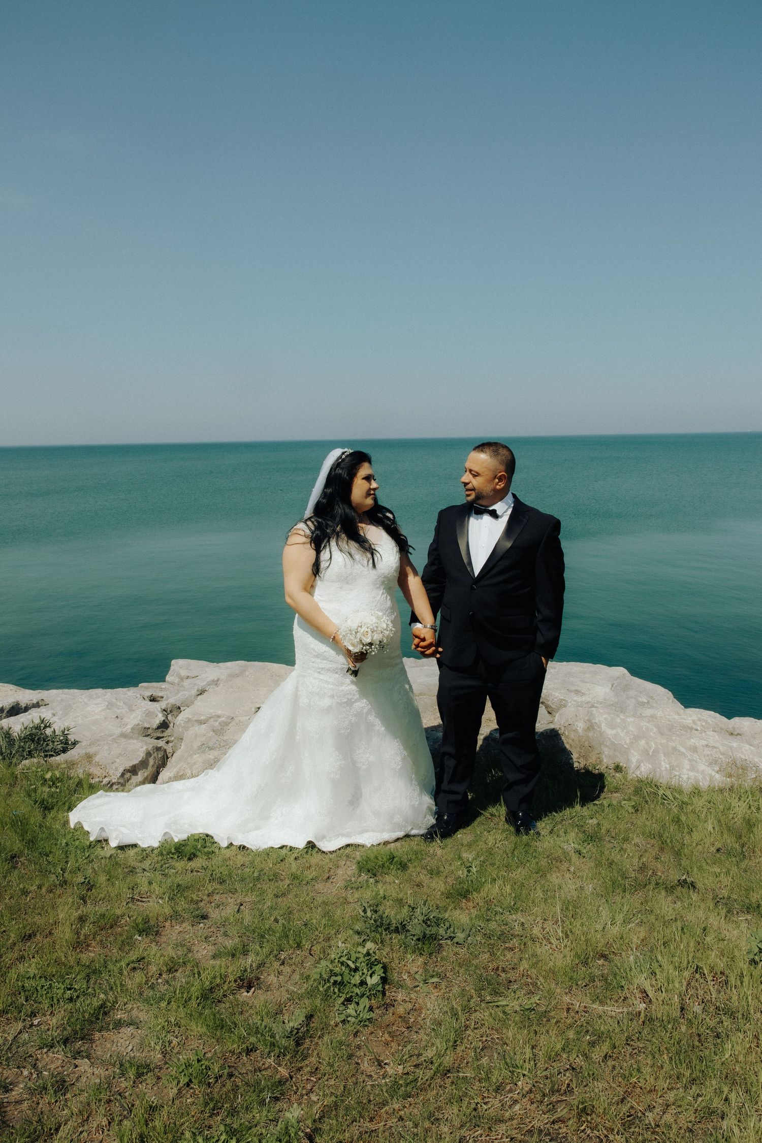 Bride and groom holding hands on rocky cliff overlooking turquoise ocean on sunny wedding day.