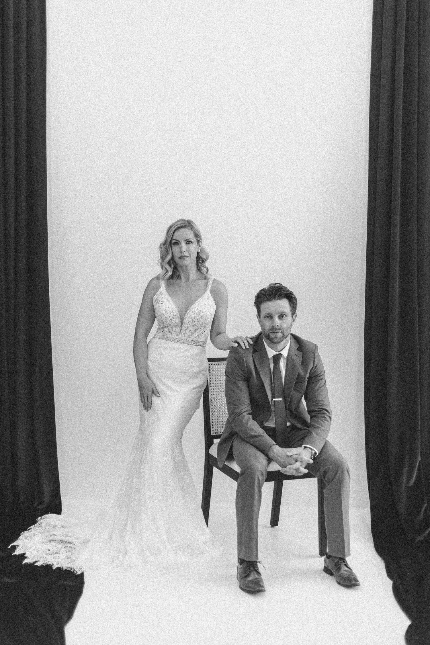 A black and white studio portrait of a couple in wedding attire, with the bride standing and groom seated on a chair.