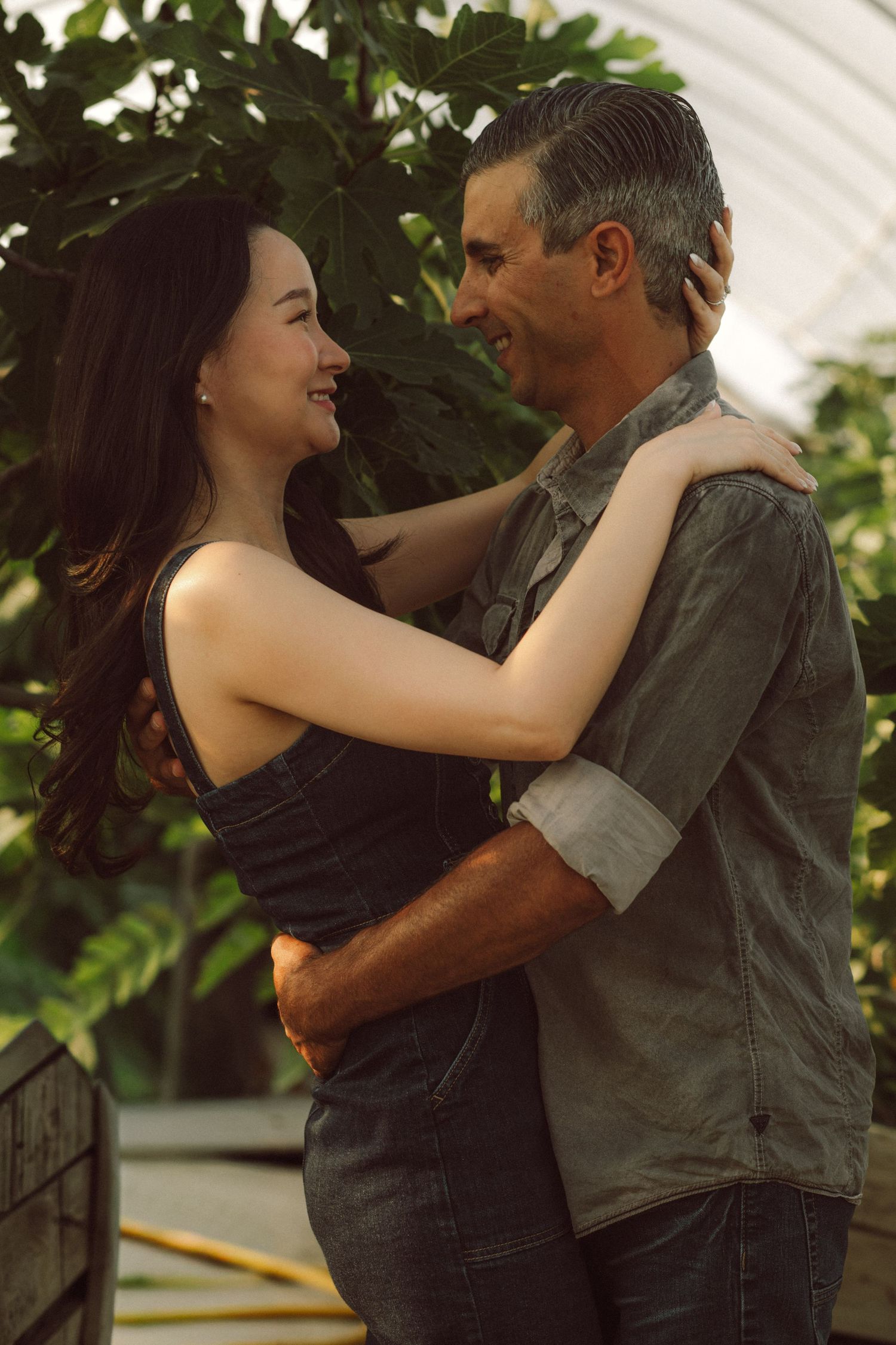 A smiling couple embraces warmly in a sunlit greenhouse surrounded by lush green plants.