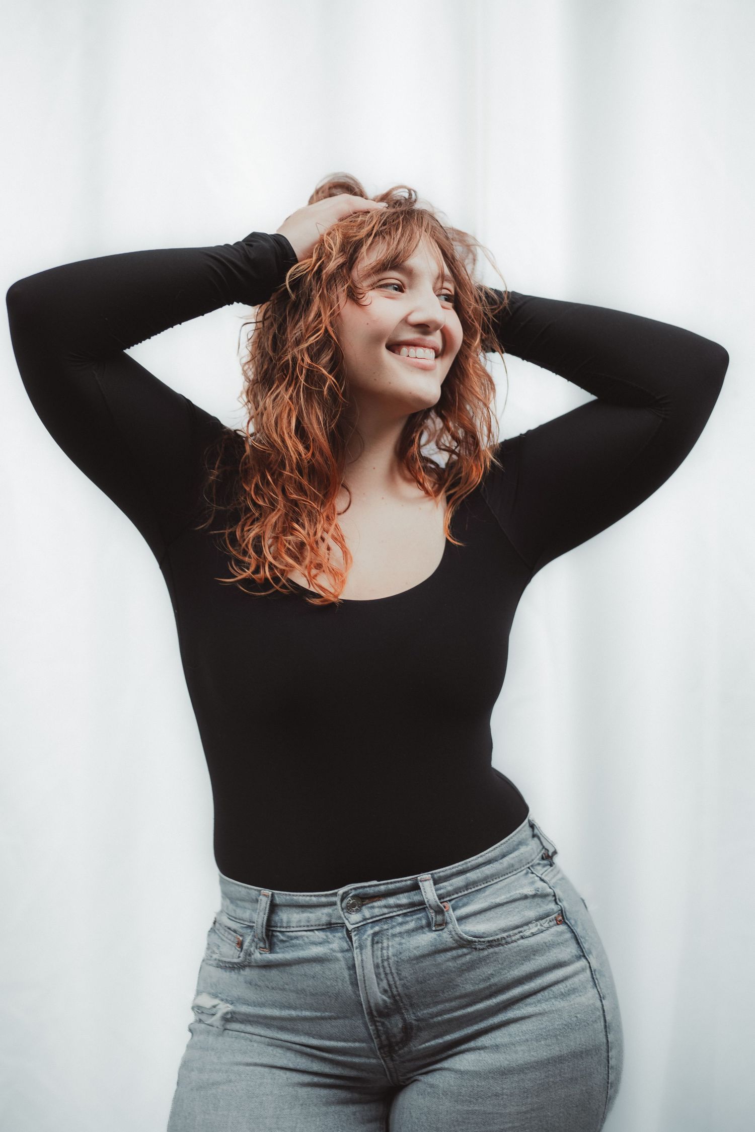 Smiling young woman with curly red hair in black long-sleeve bodysuit and jeans, posing with hands in hair.