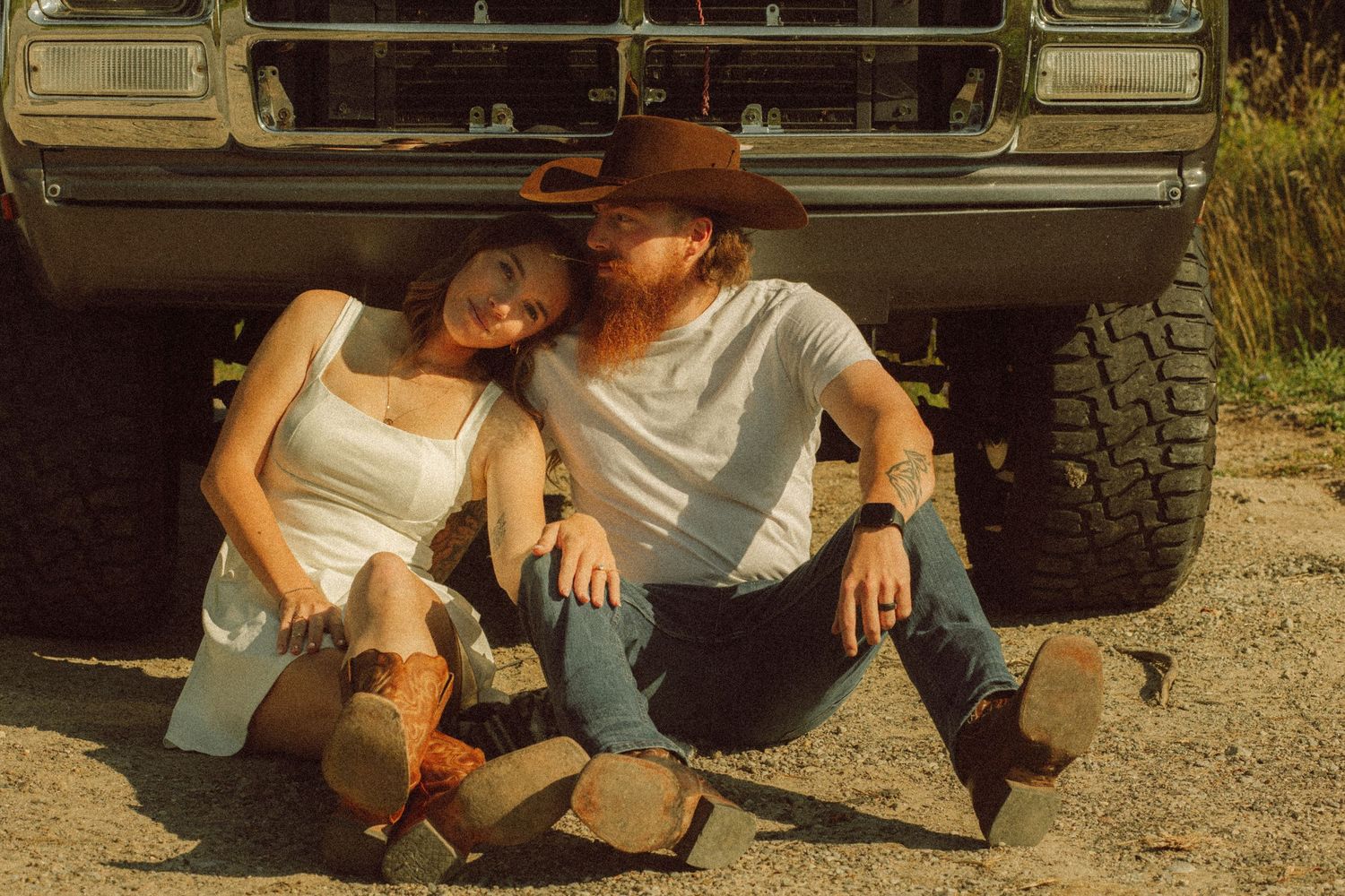 Couple in cowboy boots and hats sitting against a vintage truck on a dirt road in a rustic country setting.