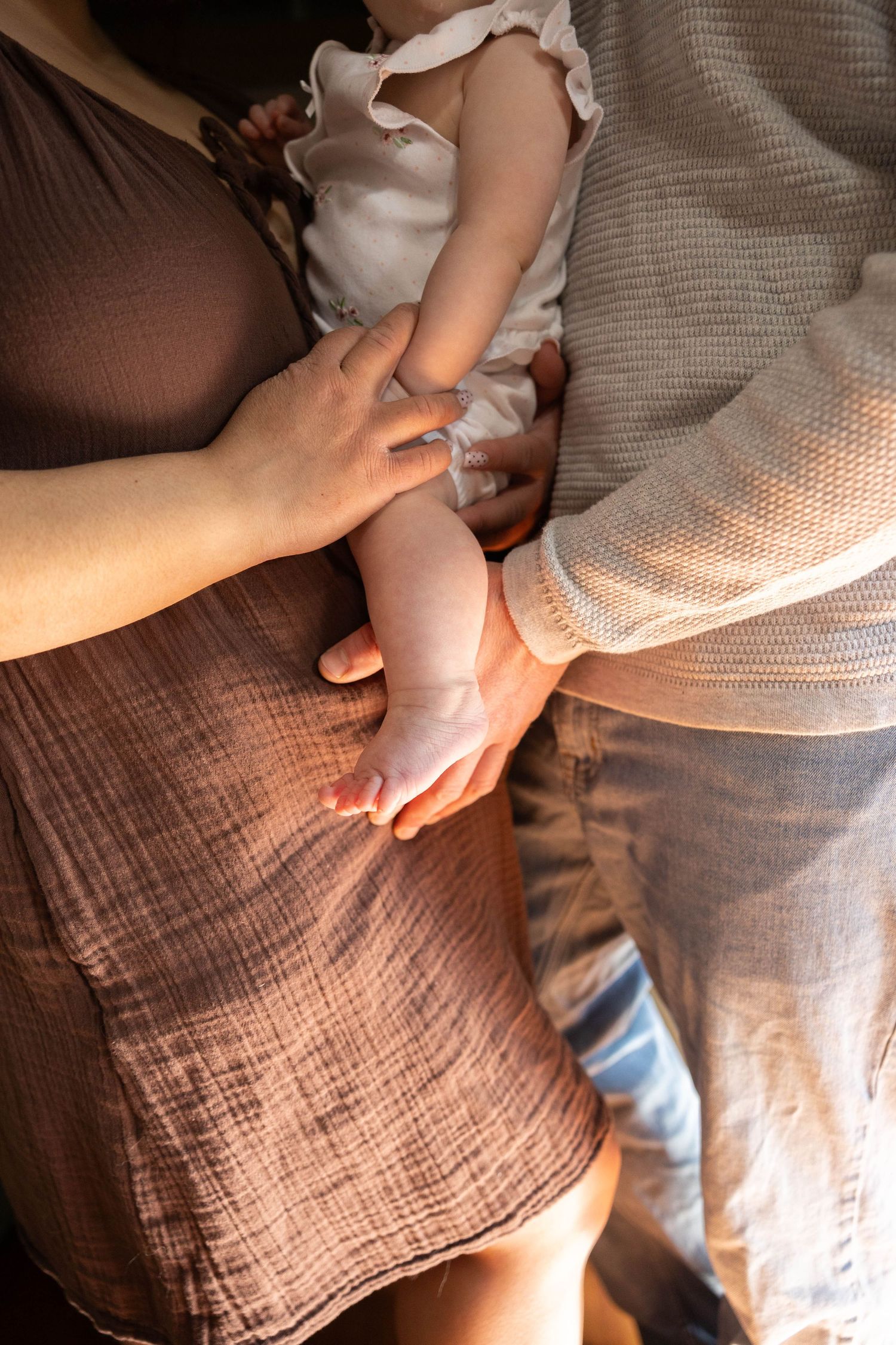 Close-up view of hands holding a baby's bare foot wearing a white dress against brown fabric.