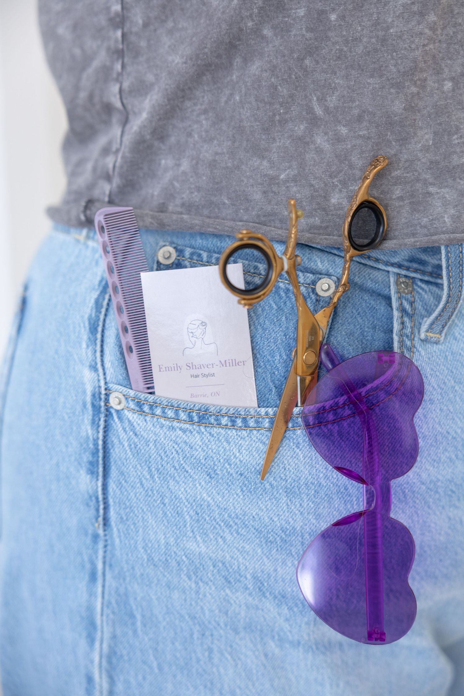 Hair stylist's jeans pocket holding gold scissors, purple comb, sunglasses, and a business card.