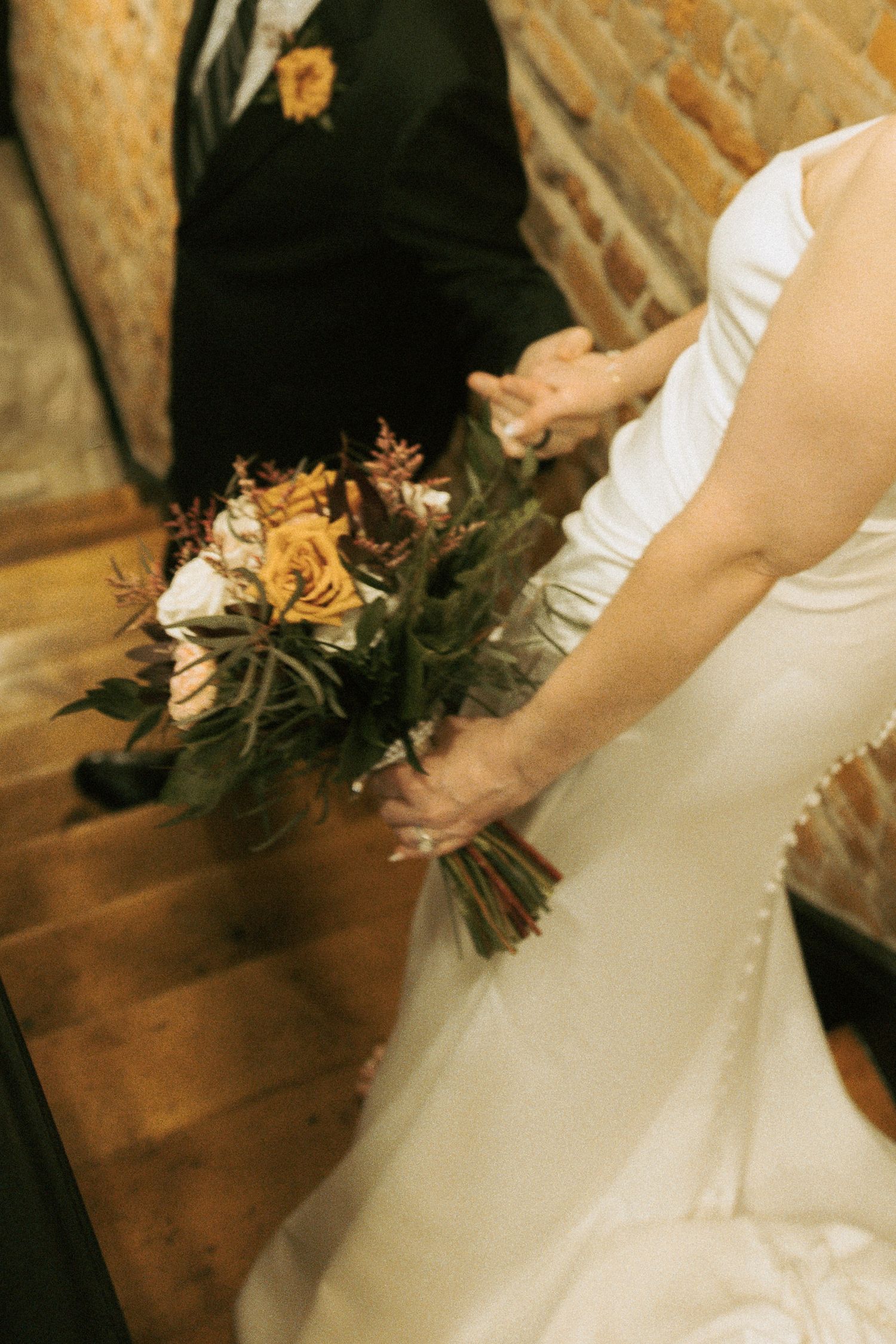Bride in white gown holding autumn bouquet with yellow roses and greenery, groom in black suit visible on stairs.