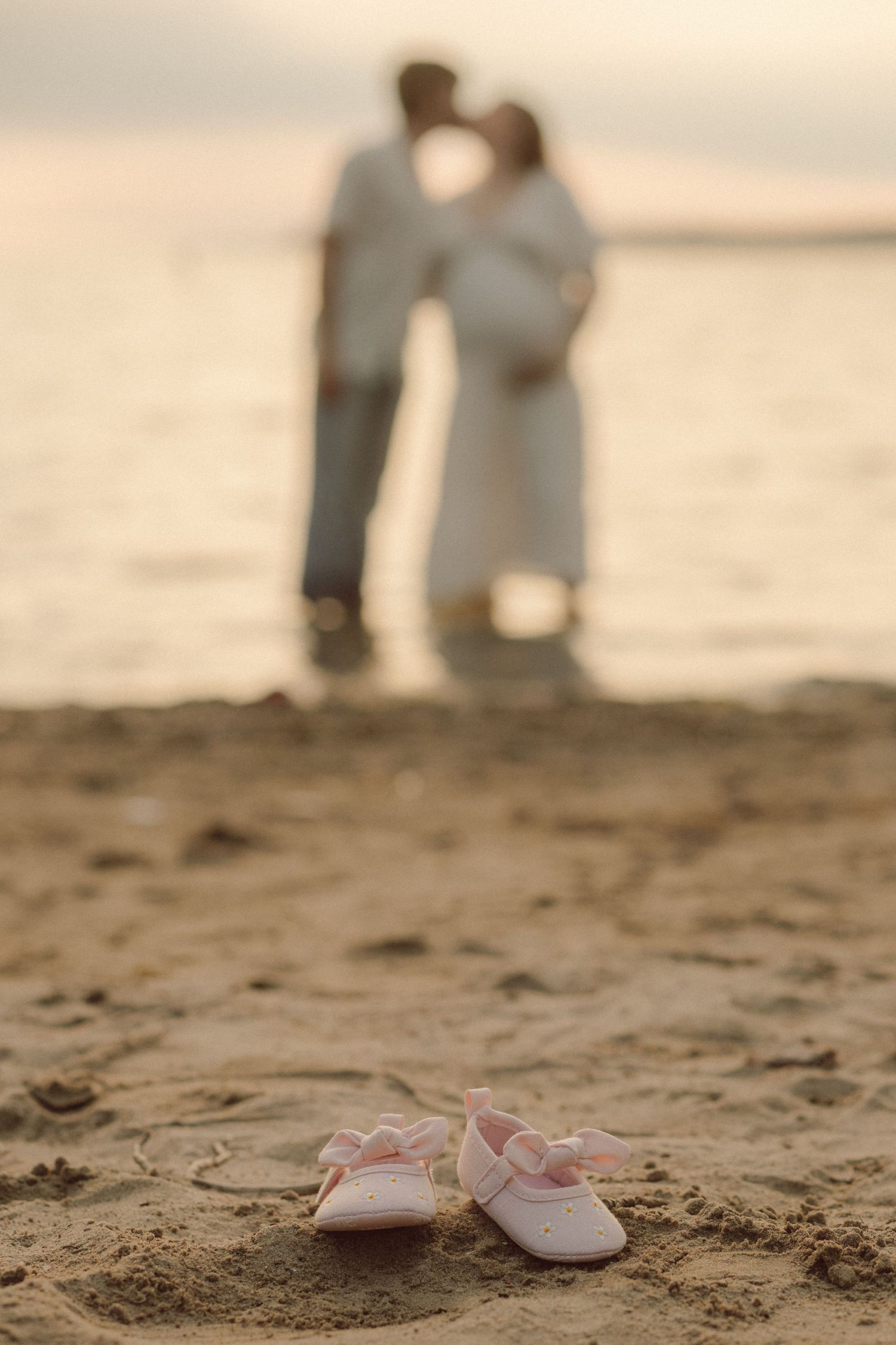 Pink baby shoes in sand with blurred pregnant couple kissing on beach at sunset.