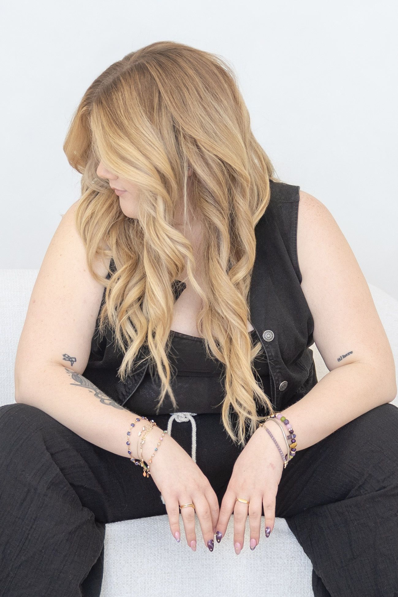 Woman with long blonde wavy hair wearing black outfit, bracelets, and rings, sitting with tattooed arms resting on lap.